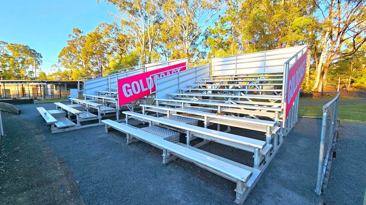 Metal bleachers with several rows of seating are on the right, featuring red signs with white text. The ground is paved with gravel. In the background, there are trees with green foliage and a building with a flat roof and fencing. The sky is clear.