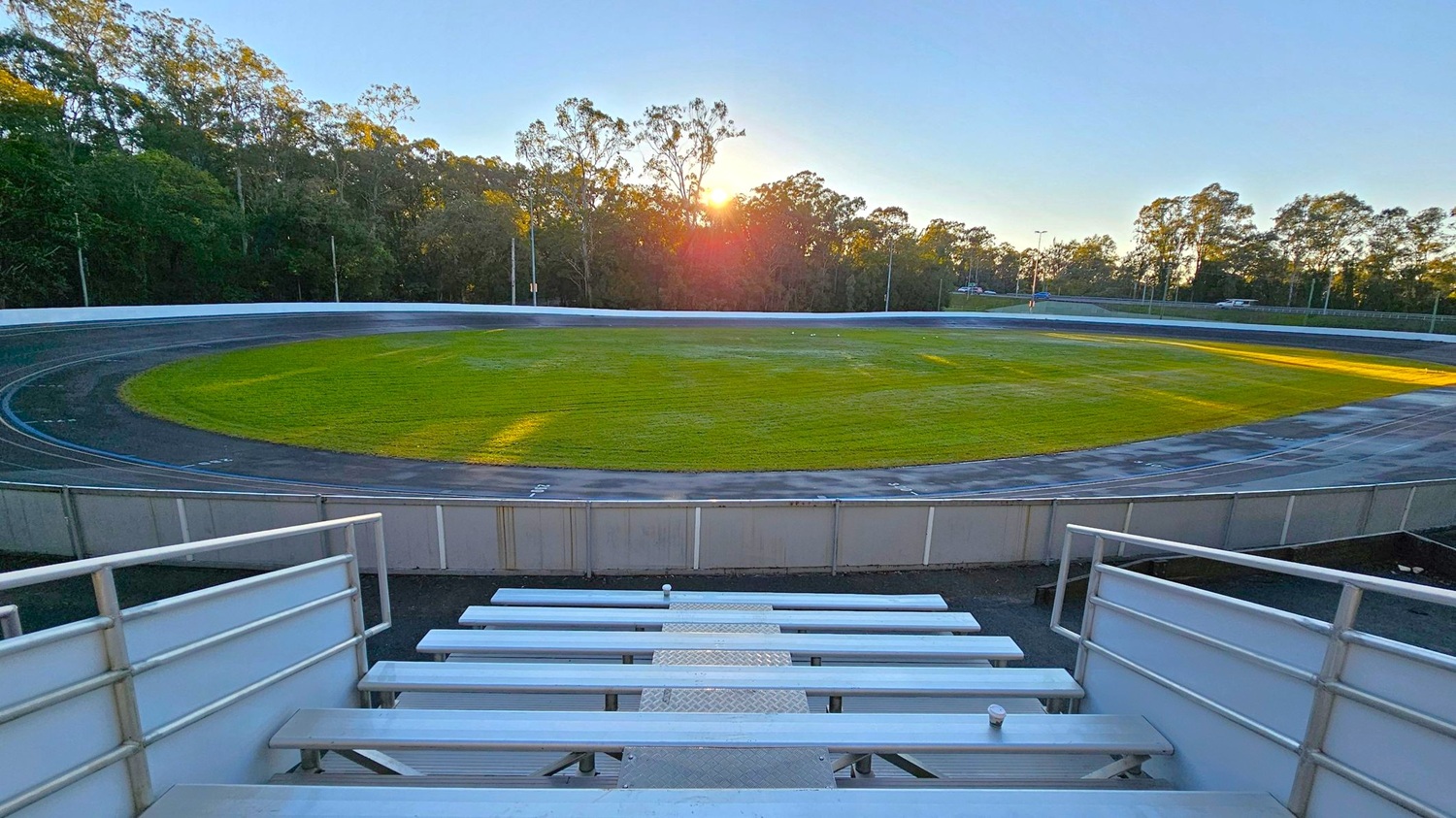 Metal bleachers in the foreground lead down to a velodrome track encircling a grassy field. The track is dark with white lines marking lanes. In the background, a line of tall trees is silhouetted against the setting sun, casting long shadows across the field. The sky is clear, and the scene is bathed in the warm glow of the sunset.