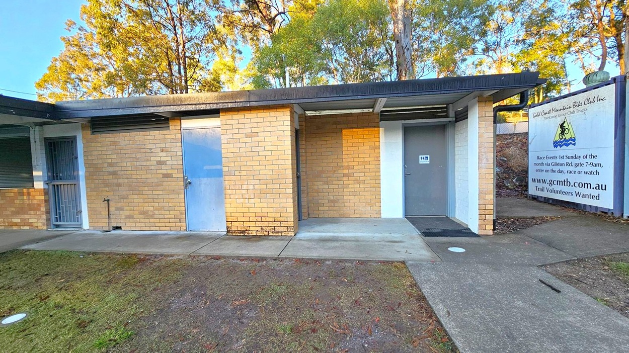 A small brick building with a flat roof. On the left, there's a metal door with a barred gate. In the centre, a solid blue door is set into the brickwork. To the right, another door with a sign above it. A large sign on the far right reads "Gold Coast Mountain Bike Club Inc." with details about race events and a website. The ground is a mix of concrete paths and patchy grass. Tall trees are visible in the background.