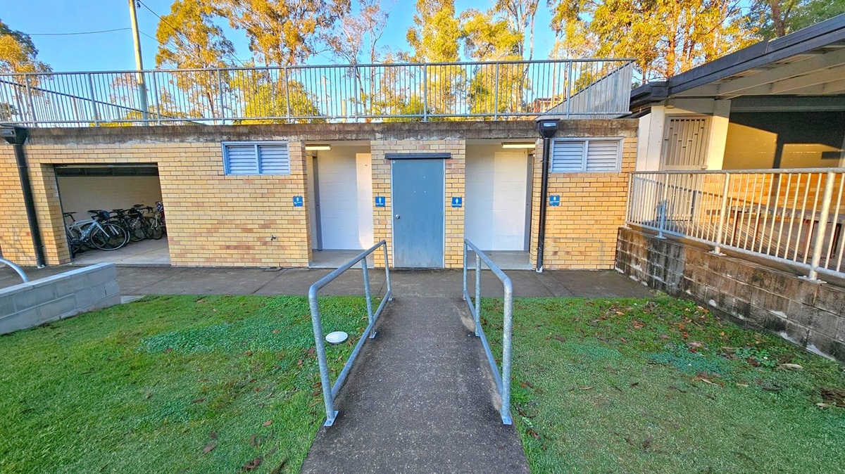 A brick building with three doors, each marked with a blue sign. The central door is closed, while the left and right doors are open, revealing small rooms. On the left, a storage area with bicycles is visible. A metal railing leads up a concrete path to the building. The foreground has a grassy area with scattered leaves. The background features trees and a clear sky. A metal fence runs along the top of the building.