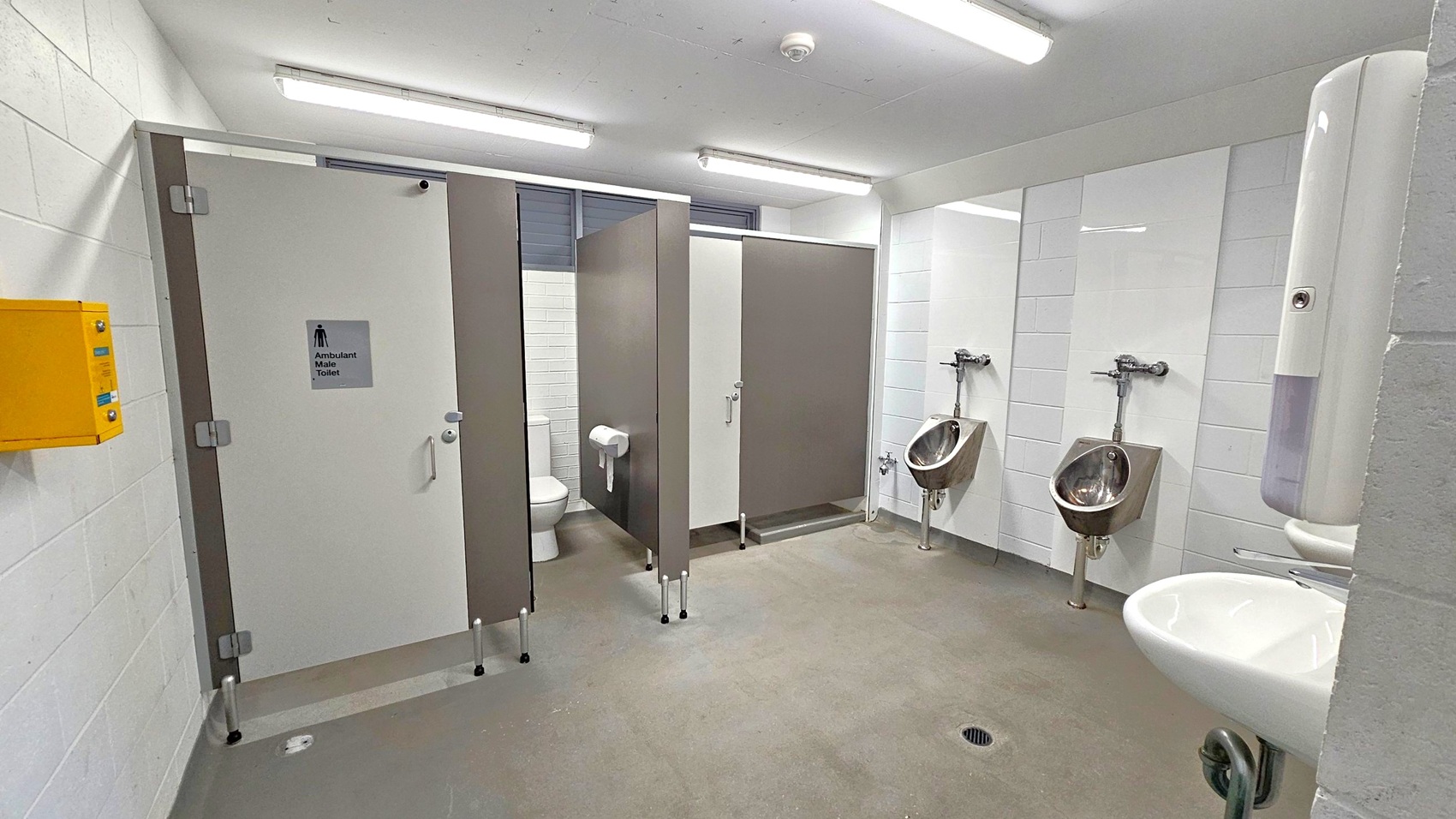 A public male toilet with a grey and white colour scheme. On the left, there is a yellow emergency box on the wall. In the centre, two toilet cubicles with grey doors, one slightly open revealing a toilet and toilet paper. On the right, two urinals are mounted on a tiled wall, with a white hand dryer above a sink. The floor is grey with a drain, and the ceiling has fluorescent lights.