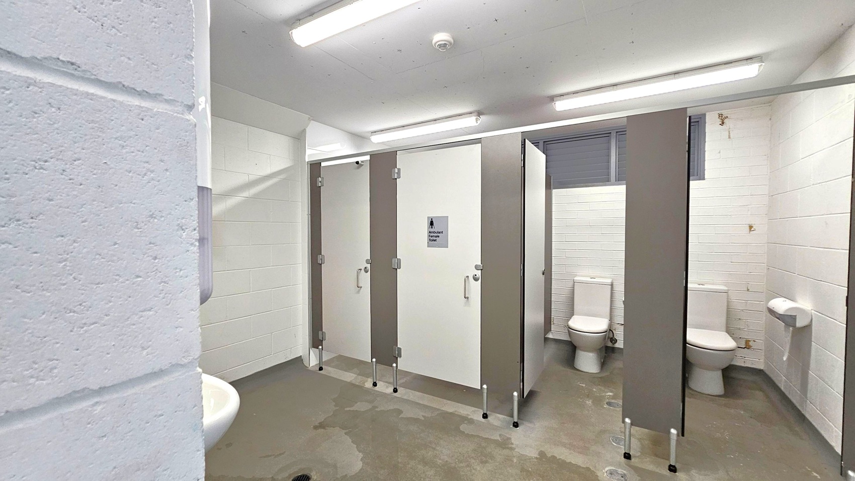 A public restroom with three stalls, each with grey and white doors. The middle stall door has a sign indicating it is an ambulant female toilet. Inside the open stall on the right, there are two white toilets. The walls are made of white brick, and the floor is a mottled green and grey. Overhead, there are fluorescent lights on the ceiling. A soap dispenser is partially visible on the left wall.