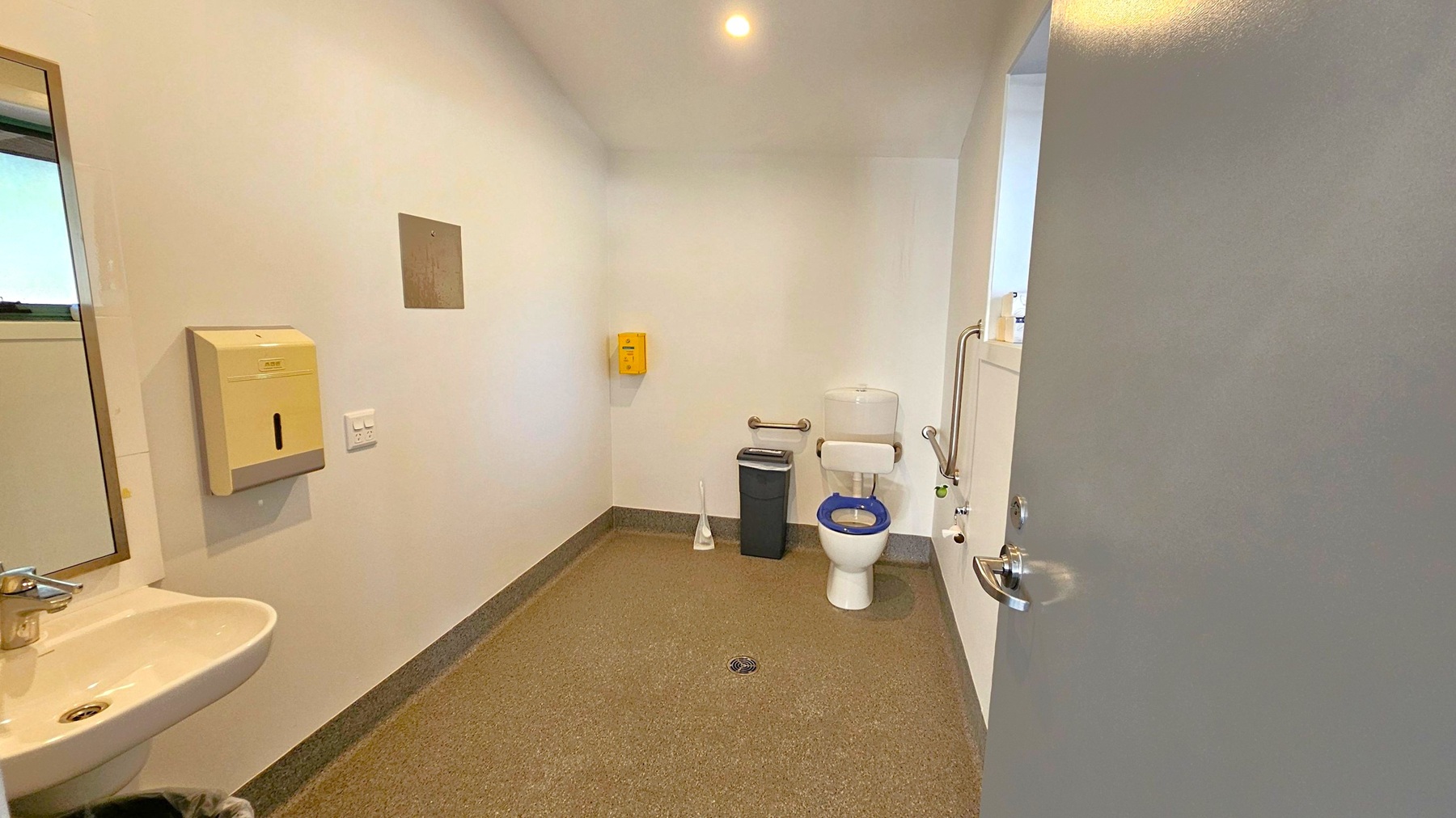 A public restroom with a grey door partially open on the right. In the centre, a toilet with a blue seat is positioned against the wall, accompanied by a grab bar and a black waste bin. A toilet brush is nearby. On the left, a small sink with a tap is mounted on the wall, above which is a soap dispenser. The floor is covered with speckled tiles, and the ceiling is plain with a single recessed light.