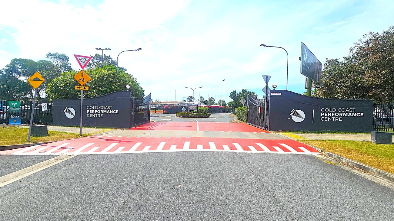 Entrance to the Gold Coast Performance Centre with black gates on both sides. Signs on the left include a "Give Way" and a pedestrian/cyclist warning. The ground features a red and white striped pedestrian crossing. Trees and bushes are visible in the background, along with a few streetlights. The road is grey, and the area is well-maintained.