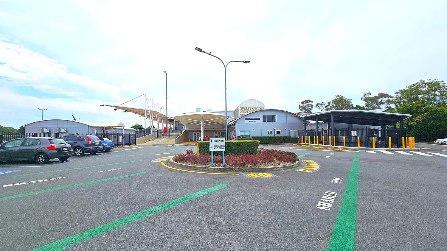 A parking area with several cars on the left. In the centre, a roundabout with a sign pointing to "Reception" and "Bus Parking & Set Down". The building in the background has a curved roof and a "Reception" sign. On the right, a covered area with yellow bollards and a pedestrian crossing. The ground has marked lanes, and there are trees in the background.