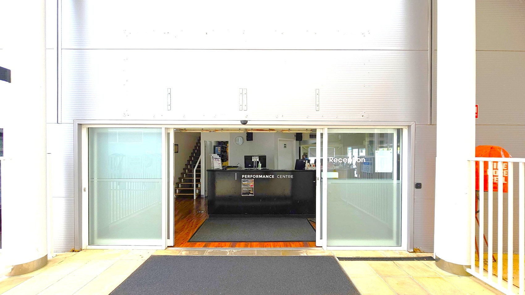A reception area with a black desk labelled "Performance Centre" in the middle. The entrance has sliding glass doors with "Reception" written on the right door. Inside, a staircase is visible to the left. The floor is tiled with a dark mat leading to the entrance. The walls are metallic with vertical lines, and there are large white columns on either side. An orange object is partially visible on the right, behind a white railing.