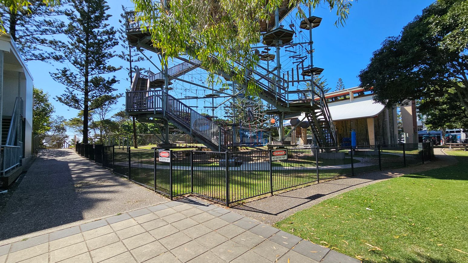 A high ropes course with multiple platforms and obstacles is enclosed by a black metal fence with "Danger No Entry" signs. On the left, a gravel path runs alongside a building, leading towards tall trees. On the right, a grassy area and a large tree provide shade. In the background, a structure with a curved roof is visible. The ground is a mix of paved tiles and grass. A high ropes course with multiple platforms and obstacles is enclosed by a black metal fence with "Danger No Entry" signs. On the left, a gravel path runs alongside a building, leading towards tall trees. On the right, a grassy area and a large tree provide shade. In the background, a structure with a curved roof is visible. The ground is a mix of paved tiles and grass.