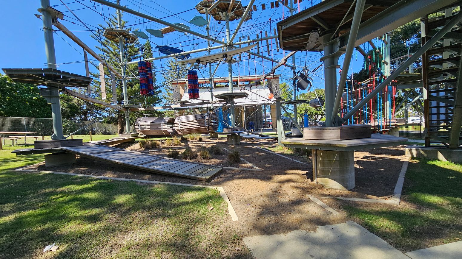 An outdoor adventure park with a high ropes course. The structure is made of metal poles and wooden platforms connected by ropes and cables. There are various obstacles, including hanging logs, rope bridges, and vertical climbing elements. The ground is a mix of grass and dirt, with some patches of sunlight and shadow. In the background, there are trees and a fence. An outdoor adventure park with a high ropes course. The structure is made of metal poles and wooden platforms connected by ropes and cables. There are various obstacles, including hanging logs, rope bridges, and vertical climbing elements. The ground is a mix of grass and dirt, with some patches of sunlight and shadow. In the background, there are trees and a fence.
