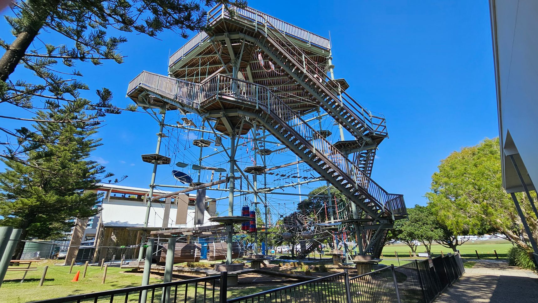 A large outdoor climbing structure with multiple levels and platforms, featuring ropes and obstacles. It is surrounded by a metal fence. On the left, there are tall trees with dense foliage. On the right, a building with a white wall is partially visible, and more trees are in the background. The ground is grassy, and a bright orange cone is placed near the structure. A large outdoor climbing structure with multiple levels and platforms, featuring ropes and obstacles. It is surrounded by a metal fence. On the left, there are tall trees with dense foliage. On the right, a building with a white wall is partially visible, and more trees are in the background. The ground is grassy, and a bright orange cone is placed near the structure.