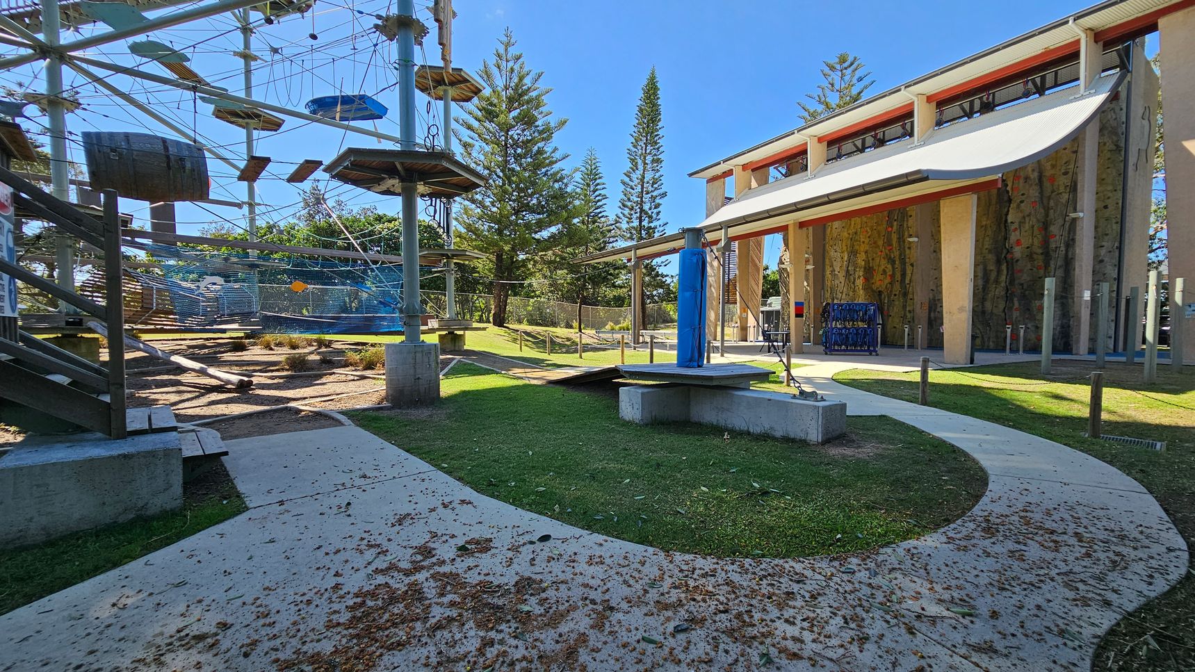 An outdoor adventure park with a high ropes course on the left, featuring platforms, ropes, and a barrel. In the centre, a curved concrete path with scattered leaves leads to a grassy area. On the right, a building with a large canopy and climbing walls. Tall trees are visible in the background. An outdoor adventure park with a high ropes course on the left, featuring platforms, ropes, and a barrel. In the centre, a curved concrete path with scattered leaves leads to a grassy area. On the right, a building with a large canopy and climbing walls. Tall trees are visible in the background.