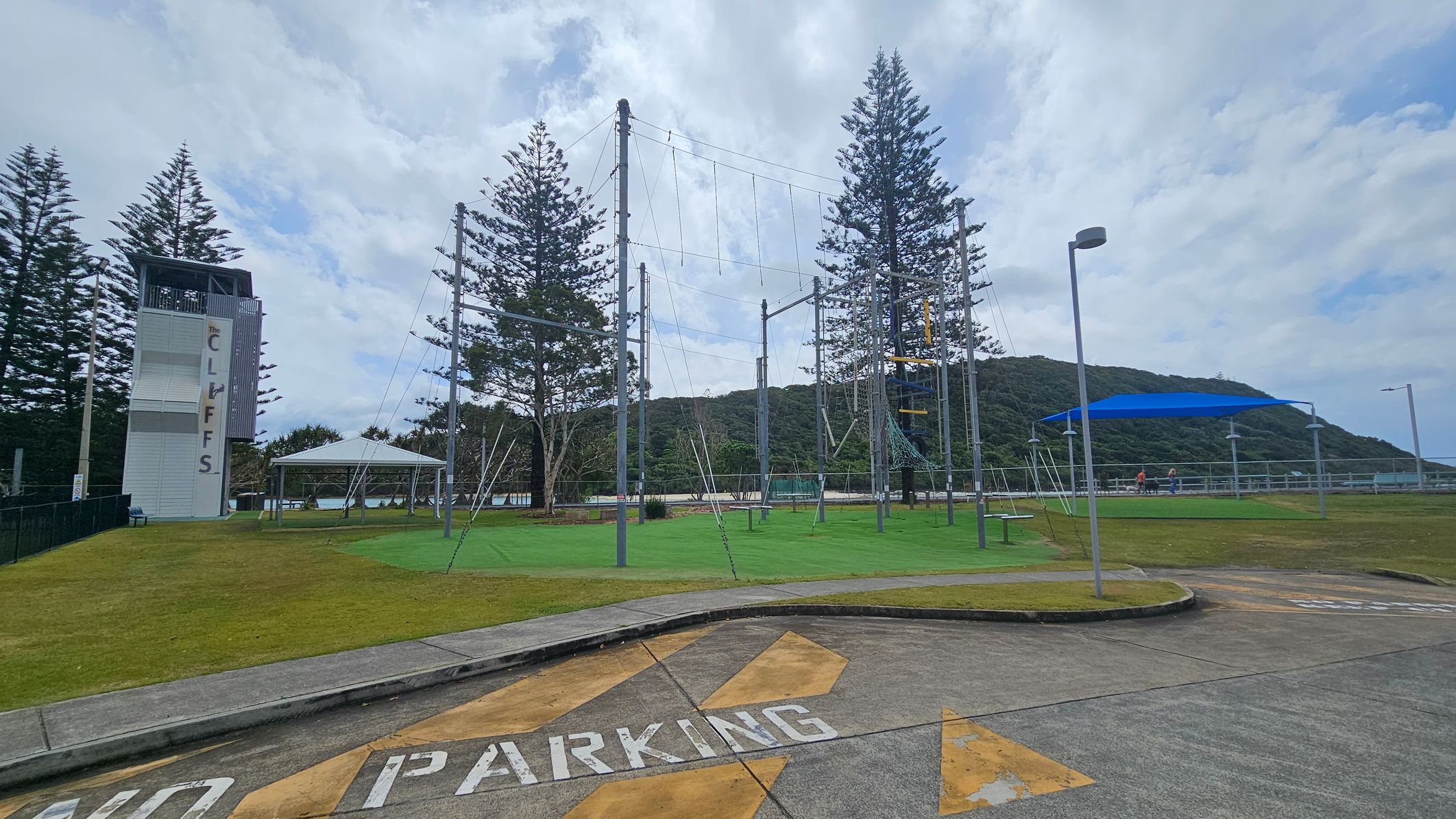 A concrete area with "NO PARKING" painted on it is in the foreground. To the left, there's a tall structure labelled "THE CLIFFS" next to a grassy area with a white canopy. In the centre, a high ropes course with metal poles and wires is visible. To the right, a blue shade structure covers a section of the grass, with a few people nearby. Tall trees and a hill are in the background. A concrete area with "NO PARKING" painted on it is in the foreground. To the left, there's a tall structure labelled "THE CLIFFS" next to a grassy area with a white canopy. In the centre, a high ropes course with metal poles and wires is visible. To the right, a blue shade structure covers a section of the grass, with a few people nearby. Tall trees and a hill are in the background.