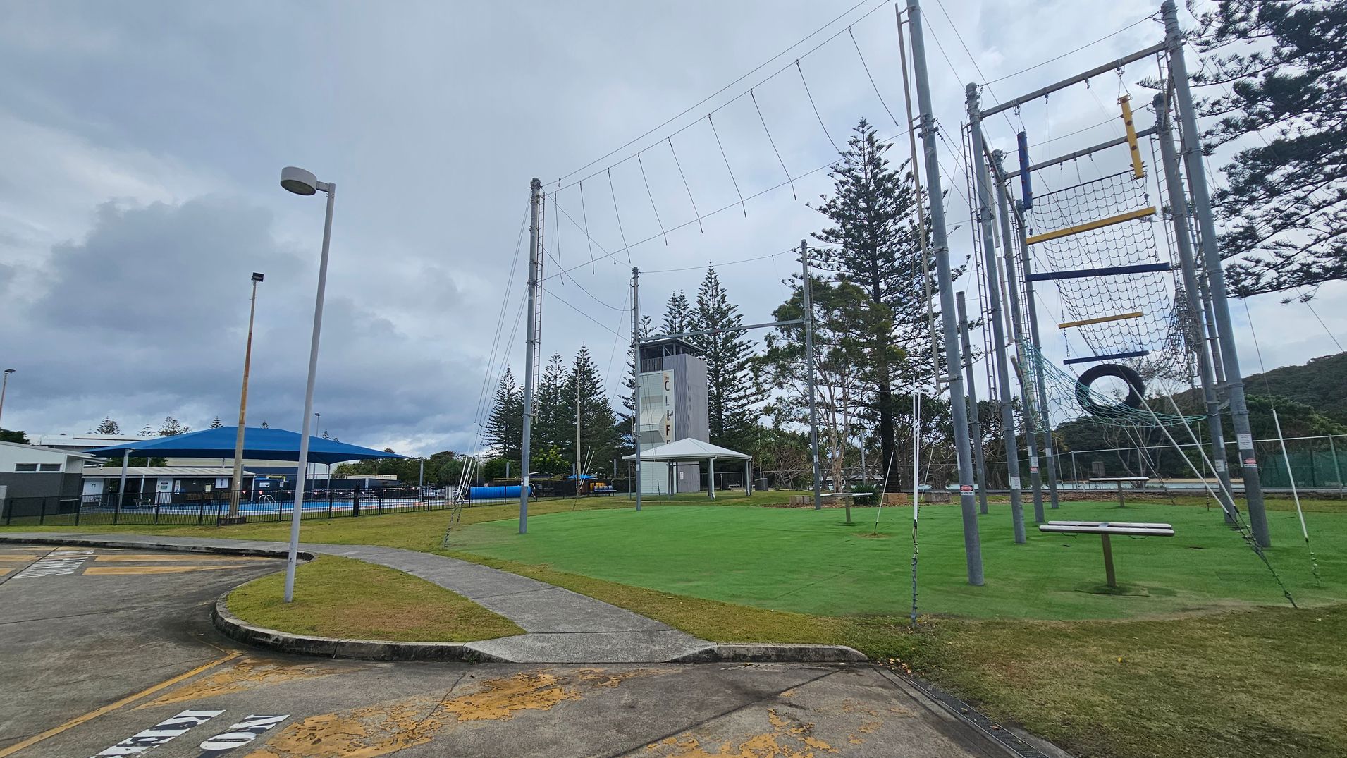 A recreational area with a high ropes course on the right, featuring nets, ropes, and a tyre. In the centre, there's a tall climbing tower with a small shelter nearby. On the left, a building with a blue roof is visible, surrounded by a black fence. The ground is a mix of concrete paths and green grass, with a few scattered trees in the background. A streetlamp stands in the foreground. A recreational area with a high ropes course on the right, featuring nets, ropes, and a tyre. In the centre, there's a tall climbing tower with a small shelter nearby. On the left, a building with a blue roof is visible, surrounded by a black fence. The ground is a mix of concrete paths and green grass, with a few scattered trees in the background. A streetlamp stands in the foreground.