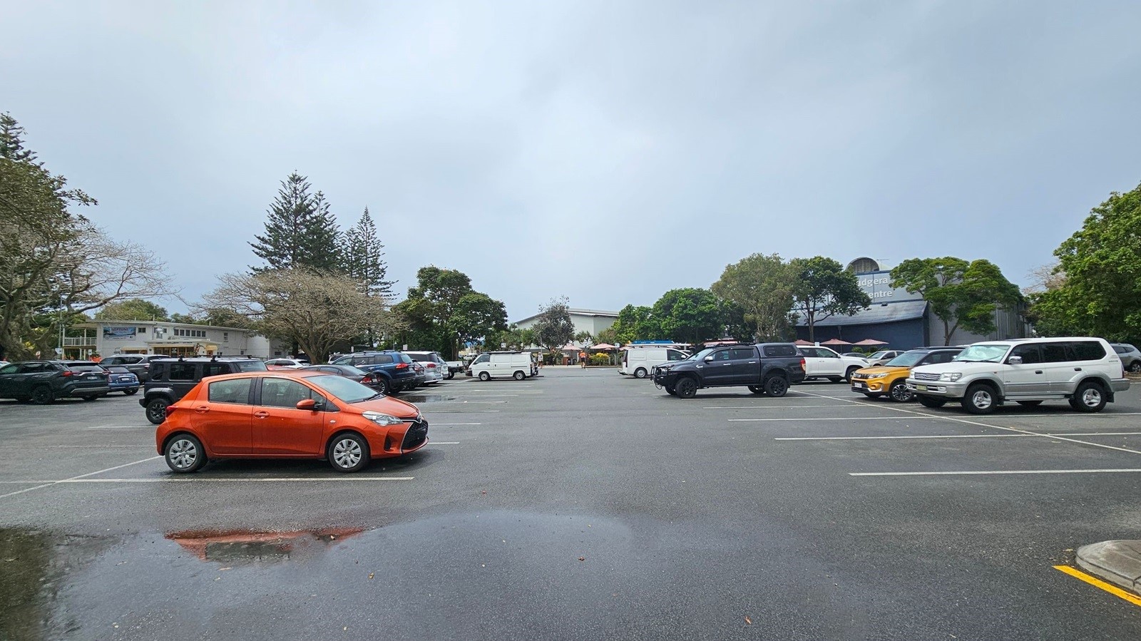 A car park with several vehicles, including an orange car prominently in the foreground. The ground is wet with a visible puddle reflecting the orange car. In the background, there are trees and a building with a sign partially visible. The area is surrounded by greenery, and the sky is overcast. A car park with several vehicles, including an orange car prominently in the foreground. The ground is wet with a visible puddle reflecting the orange car. In the background, there are trees and a building with a sign partially visible. The area is surrounded by greenery, and the sky is overcast.