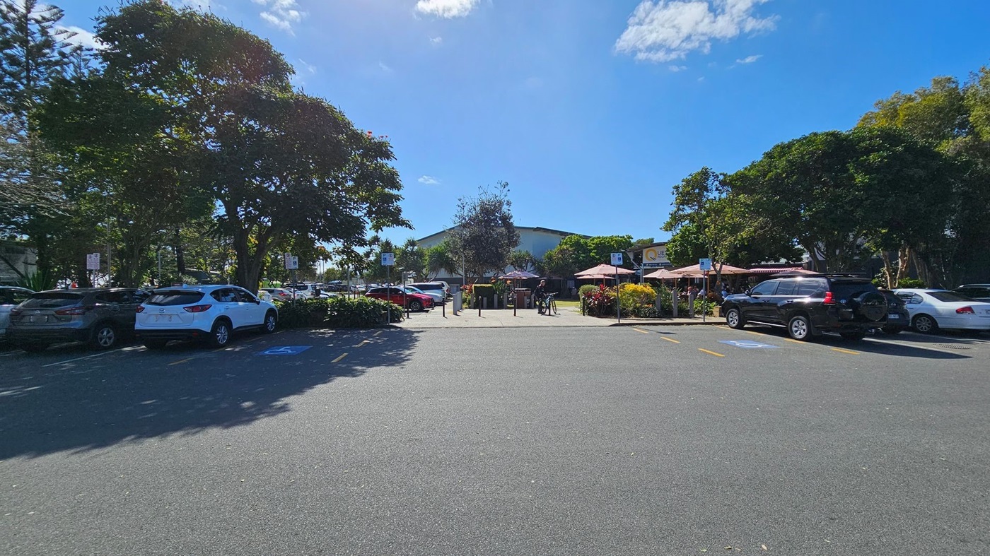 A car park with several vehicles, including a white SUV and a black SUV, is surrounded by trees and greenery. In the background, there are outdoor seating areas with umbrellas and a building with a sign that reads "QUE." A person is standing near a bicycle. The ground is paved, and there are marked parking spaces, including a disabled parking spot. A car park with several vehicles, including a white SUV and a black SUV, is surrounded by trees and greenery. In the background, there are outdoor seating areas with umbrellas and a building with a sign that reads "QUE." A person is standing near a bicycle. The ground is paved, and there are marked parking spaces, including a disabled parking spot.