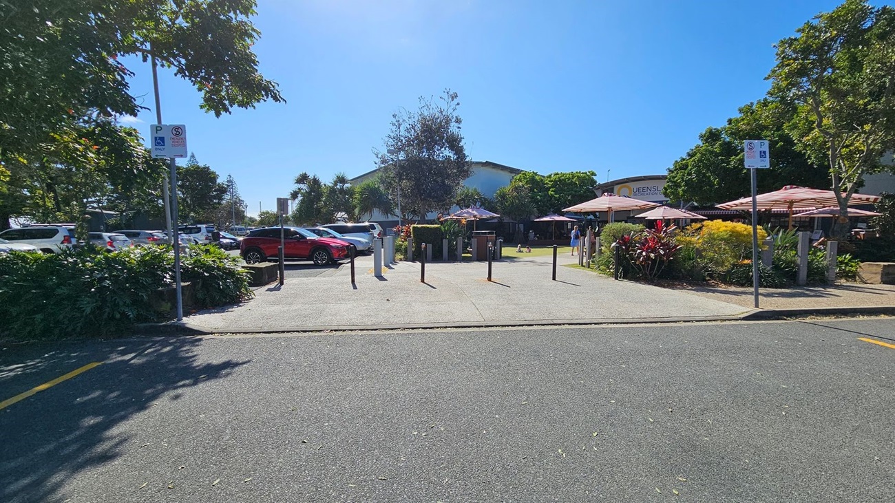 A car park with several vehicles, including a red car on the left. In the centre, there are bollards and a pathway leading to a building partially visible in the background. On the right, there are outdoor seating areas with red and white striped umbrellas, surrounded by greenery and colourful plants. Parking signs are visible on both sides. Trees provide shade throughout the area. A car park with several vehicles, including a red car on the left. In the centre, there are bollards and a pathway leading to a building partially visible in the background. On the right, there are outdoor seating areas with red and white striped umbrellas, surrounded by greenery and colourful plants. Parking signs are visible on both sides. Trees provide shade throughout the area.