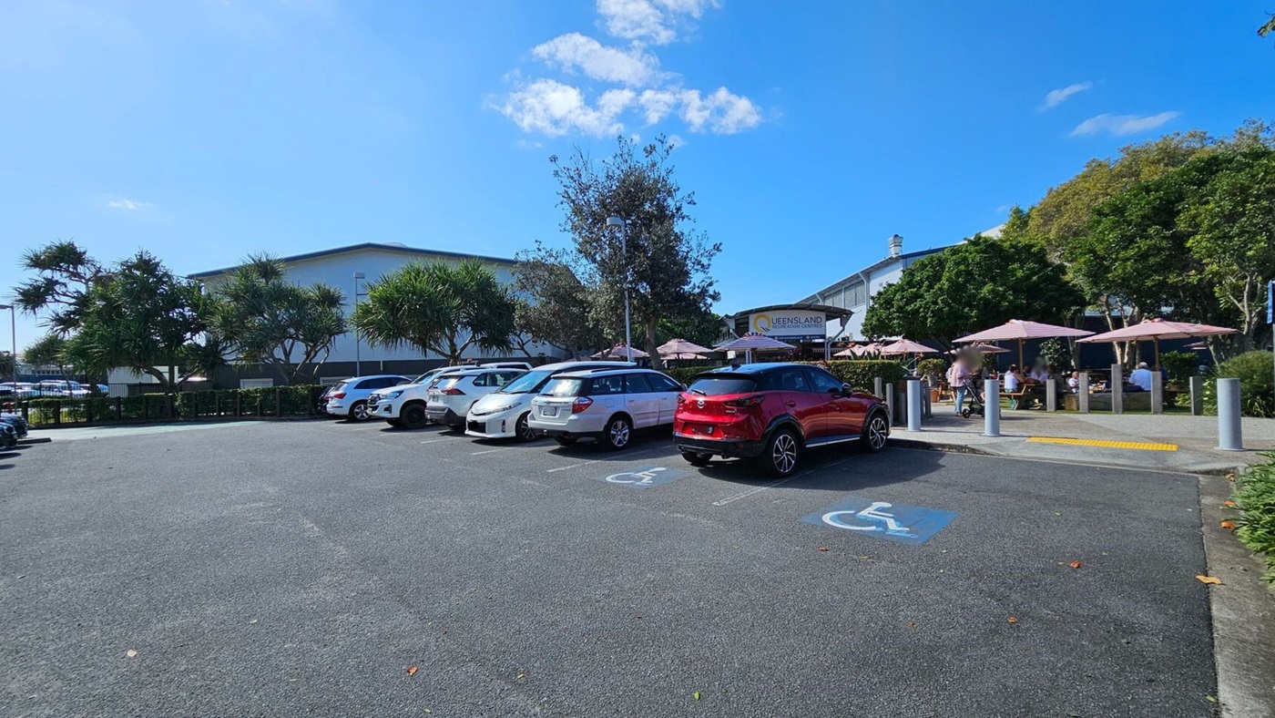 A car park with several vehicles, including a red car in a disabled parking space. On the right, there are outdoor tables with umbrellas and people seated, likely a café or restaurant area. A sign reads "Queensland" in the background. Trees and shrubs are scattered around, providing some greenery. The ground is asphalt, and there are bollards near the seating area. A car park with several vehicles, including a red car in a disabled parking space. On the right, there are outdoor tables with umbrellas and people seated, likely a café or restaurant area. A sign reads "Queensland" in the background. Trees and shrubs are scattered around, providing some greenery. The ground is asphalt, and there are bollards near the seating area.