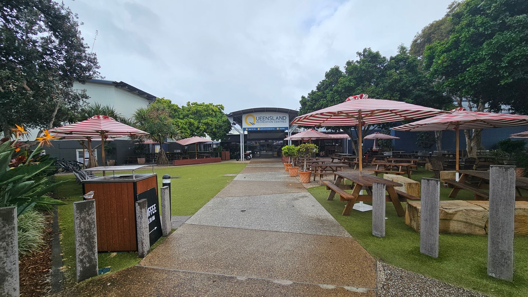 A pathway leads to a building with a sign reading "Queensland Recreation Centres" in the background. On both sides of the path are picnic tables with red and white striped umbrellas. The area is surrounded by lush green trees and plants, including bird of paradise flowers on the left. The ground is a mix of grass and concrete, and there are several concrete bollards along the path. A pathway leads to a building with a sign reading "Queensland Recreation Centres" in the background. On both sides of the path are picnic tables with red and white striped umbrellas. The area is surrounded by lush green trees and plants, including bird of paradise flowers on the left. The ground is a mix of grass and concrete, and there are several concrete bollards along the path.