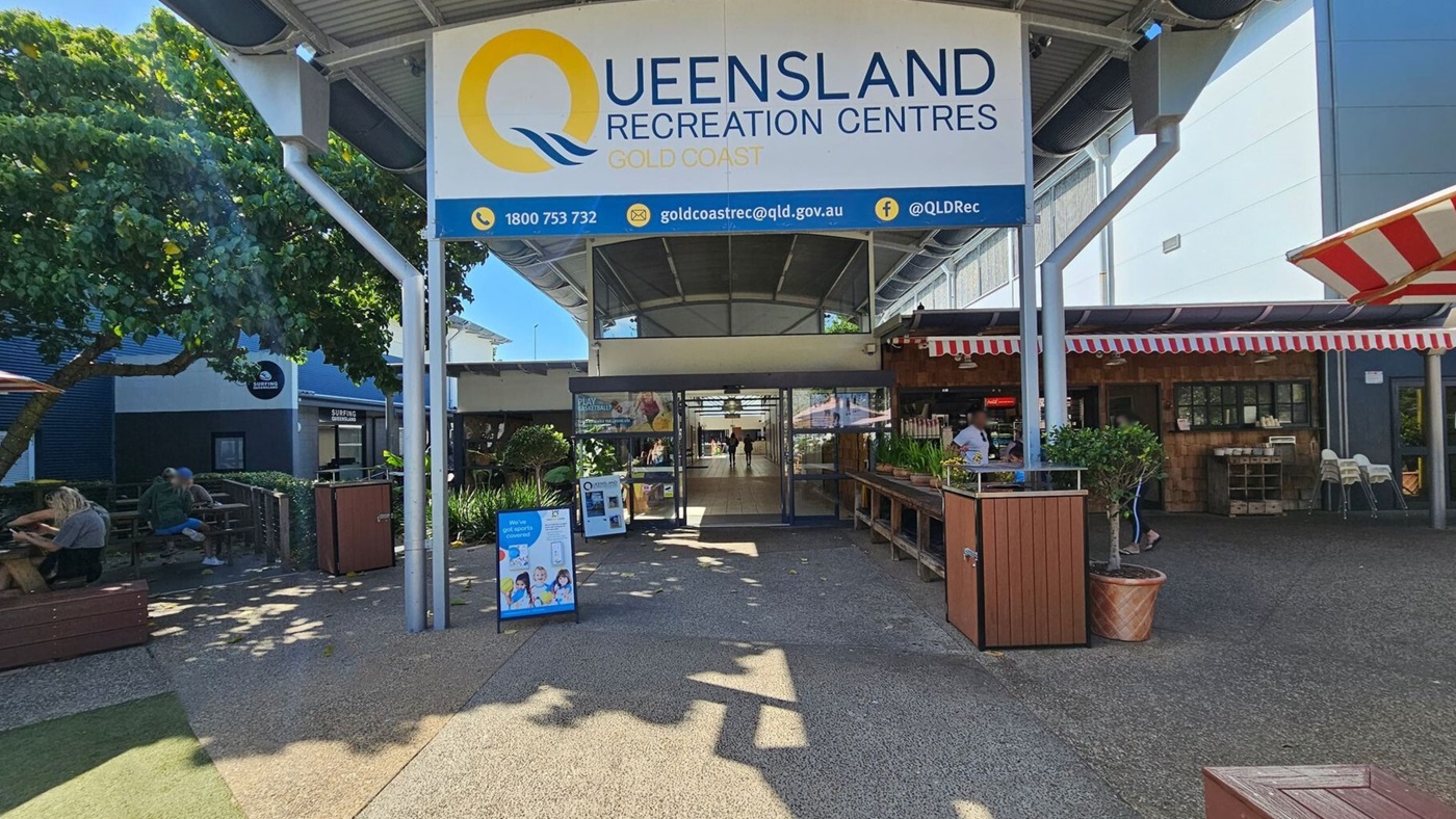 Entrance to Queensland Recreation Centres, Gold Coast. A large sign is overhead. On the left, people sit at wooden benches under a tree. On the right, a café with a red and white striped awning and a counter with plants. The centre has glass doors leading inside. The ground is paved, and the ceiling is metallic with beams. Entrance to Queensland Recreation Centres, Gold Coast. A large sign is overhead. On the left, people sit at wooden benches under a tree. On the right, a café with a red and white striped awning and a counter with plants. The centre has glass doors leading inside. The ground is paved, and the ceiling is metallic with beams.