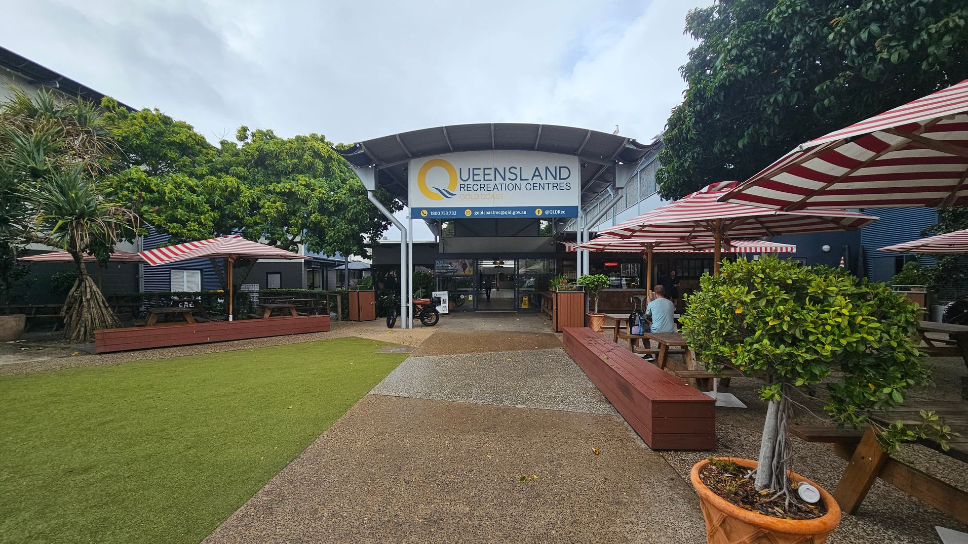 A courtyard with a pathway leading to the entrance of the Queensland Recreation Centres. The building has a large sign above the entrance. On the left, there are trees and picnic tables under red and white striped umbrellas. On the right, more picnic tables and benches are shaded by similar umbrellas. A potted tree is in the foreground on the right. The ground is a mix of grass and paved areas. A courtyard with a pathway leading to the entrance of the Queensland Recreation Centres. The building has a large sign above the entrance. On the left, there are trees and picnic tables under red and white striped umbrellas. On the right, more picnic tables and benches are shaded by similar umbrellas. A potted tree is in the foreground on the right. The ground is a mix of grass and paved areas.