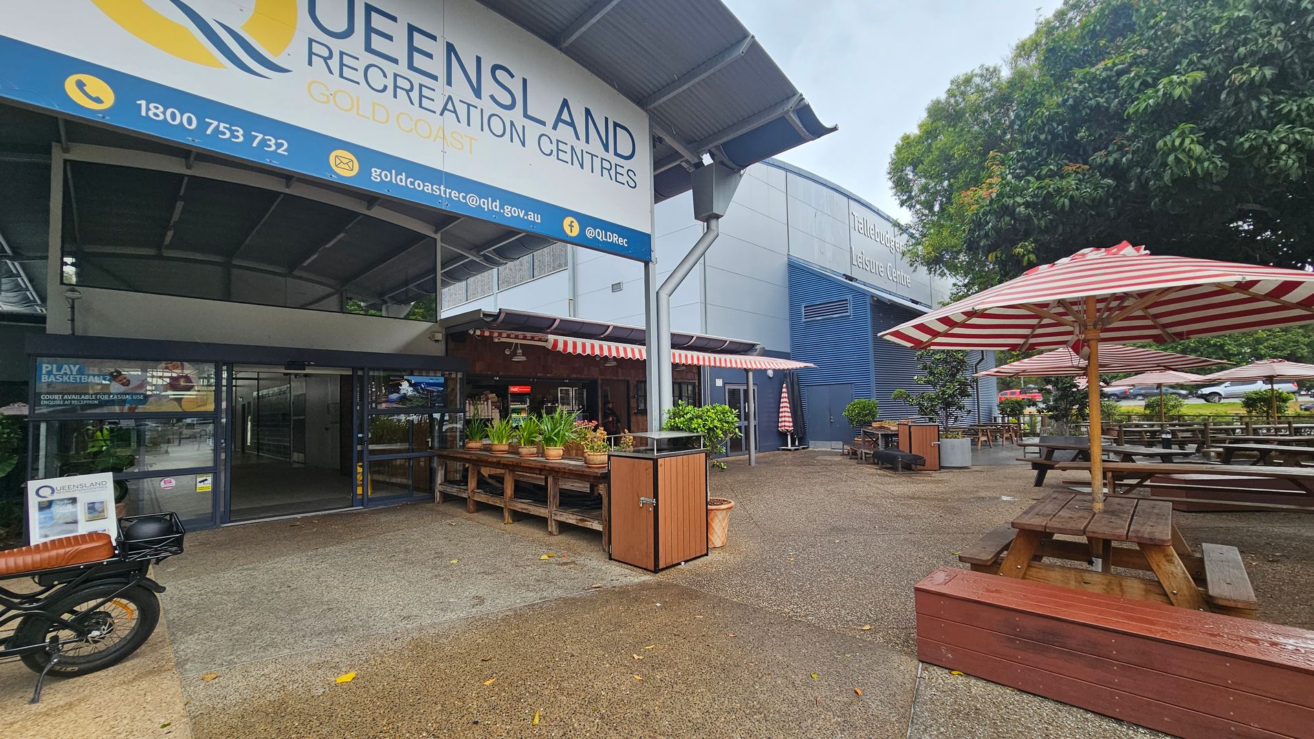 Entrance to Queensland Recreation Centres Gold Coast with a large sign above. On the left, a motorbike is parked near the entrance. In the centre, there are potted plants on wooden tables under a canopy with red and white striped awnings. On the right, picnic tables with matching striped umbrellas are set on a paved area. A large tree provides shade in the background, and a building labelled "Tallebudgera Leisure Centre" is visible. Entrance to Queensland Recreation Centres Gold Coast with a large sign above. On the left, a motorbike is parked near the entrance. In the centre, there are potted plants on wooden tables under a canopy with red and white striped awnings. On the right, picnic tables with matching striped umbrellas are set on a paved area. A large tree provides shade in the background, and a building labelled "Tallebudgera Leisure Centre" is visible.