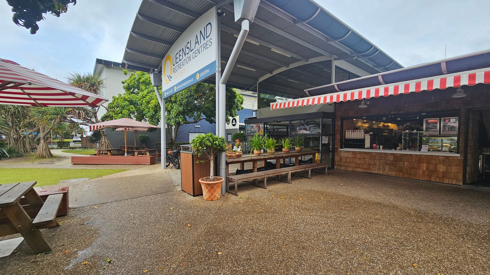 Outdoor seating area with wooden picnic tables and benches on the left, under red and white striped umbrellas. In the centre, a sign reads "Queensland Recreation Centres" above a row of potted plants on a wooden table. On the right, a café with a red and white striped awning displays a counter with food and drinks inside. The ground is a mix of grass and textured pavement, and there are trees in the background. Outdoor seating area with wooden picnic tables and benches on the left, under red and white striped umbrellas. In the centre, a sign reads "Queensland Recreation Centres" above a row of potted plants on a wooden table. On the right, a café with a red and white striped awning displays a counter with food and drinks inside. The ground is a mix of grass and textured pavement, and there are trees in the background.