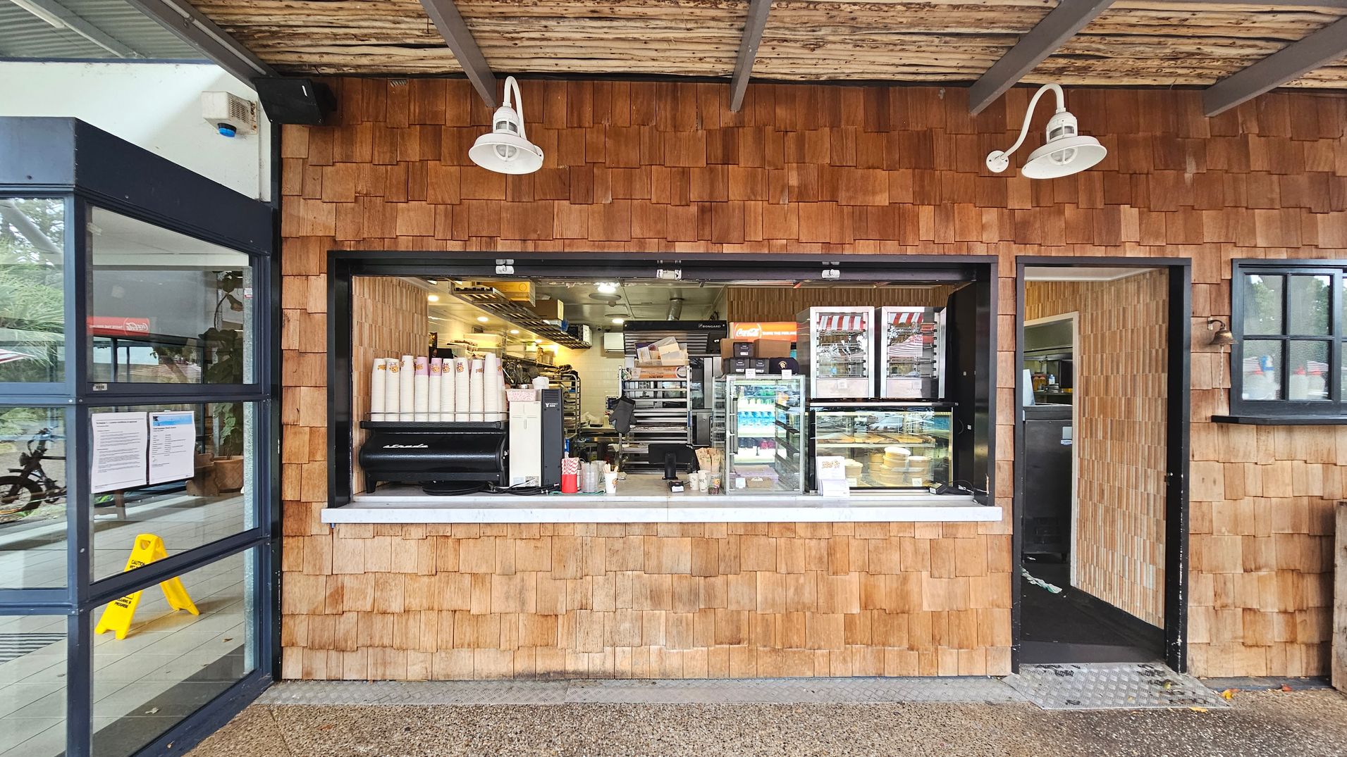 A small café with a wooden shingle exterior. In the centre, there's a service window displaying coffee cups, a coffee machine, and a glass case with pastries. To the right, an open door leads inside. Above, two white lamps are mounted on the wall. On the left, a glass door with notices and a yellow caution sign is visible. The ceiling is made of wooden slats, and the floor is a textured concrete surface. A small café with a wooden shingle exterior. In the centre, there's a service window displaying coffee cups, a coffee machine, and a glass case with pastries. To the right, an open door leads inside. Above, two white lamps are mounted on the wall. On the left, a glass door with notices and a yellow caution sign is visible. The ceiling is made of wooden slats, and the floor is a textured concrete surface.
