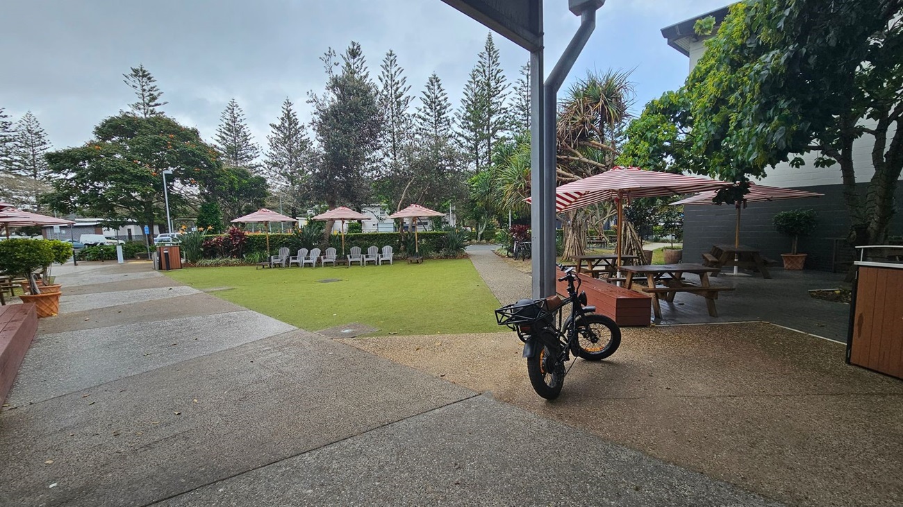 A courtyard with a mix of concrete and grass surfaces. On the left, there are several picnic tables with red and white striped umbrellas. In the centre, a grassy area is surrounded by white chairs and more umbrellas. On the right, a bicycle is parked near a wooden bench. Trees and shrubs are scattered throughout, providing shade and greenery. A building with a dark wall is partially visible on the right. A courtyard with a mix of concrete and grass surfaces. On the left, there are several picnic tables with red and white striped umbrellas. In the centre, a grassy area is surrounded by white chairs and more umbrellas. On the right, a bicycle is parked near a wooden bench. Trees and shrubs are scattered throughout, providing shade and greenery. A building with a dark wall is partially visible on the right.