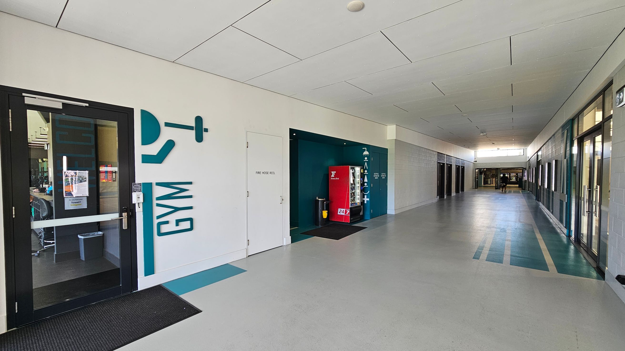 A corridor with a light grey floor and a white ceiling. On the left, there's a glass door with "GYM" written beside it, leading to a gym area. Next to it is a door labelled "FIRE HOSE REEL". Further along, there's a red vending machine in a recessed area. On the right, there are several glass doors and windows. In the background, two people are walking towards the end of the corridor, where more doors and windows are visible. A corridor with a light grey floor and a white ceiling. On the left, there's a glass door with "GYM" written beside it, leading to a gym area. Next to it is a door labelled "FIRE HOSE REEL". Further along, there's a red vending machine in a recessed area. On the right, there are several glass doors and windows. In the background, two people are walking towards the end of the corridor, where more doors and windows are visible.