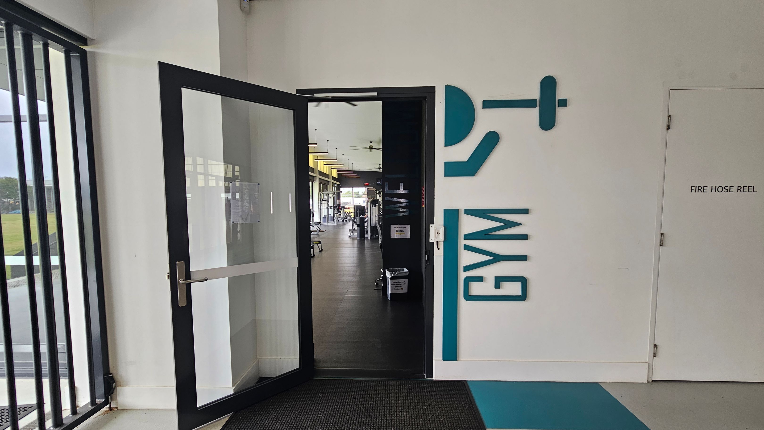 A corridor with a light grey floor and a white ceiling. On the left, there's a glass door with "GYM" written beside it, leading to a gym area. Next to it is a door labelled "FIRE HOSE REEL". Further along, there's a red vending machine in a recessed area. On the right, there are several glass doors and windows. In the background, two people are walking towards the end of the corridor, where more doors and windows are visible. A corridor with a light grey floor and a white ceiling. On the left, there's a glass door with "GYM" written beside it, leading to a gym area. Next to it is a door labelled "FIRE HOSE REEL". Further along, there's a red vending machine in a recessed area. On the right, there are several glass doors and windows. In the background, two people are walking towards the end of the corridor, where more doors and windows are visible.