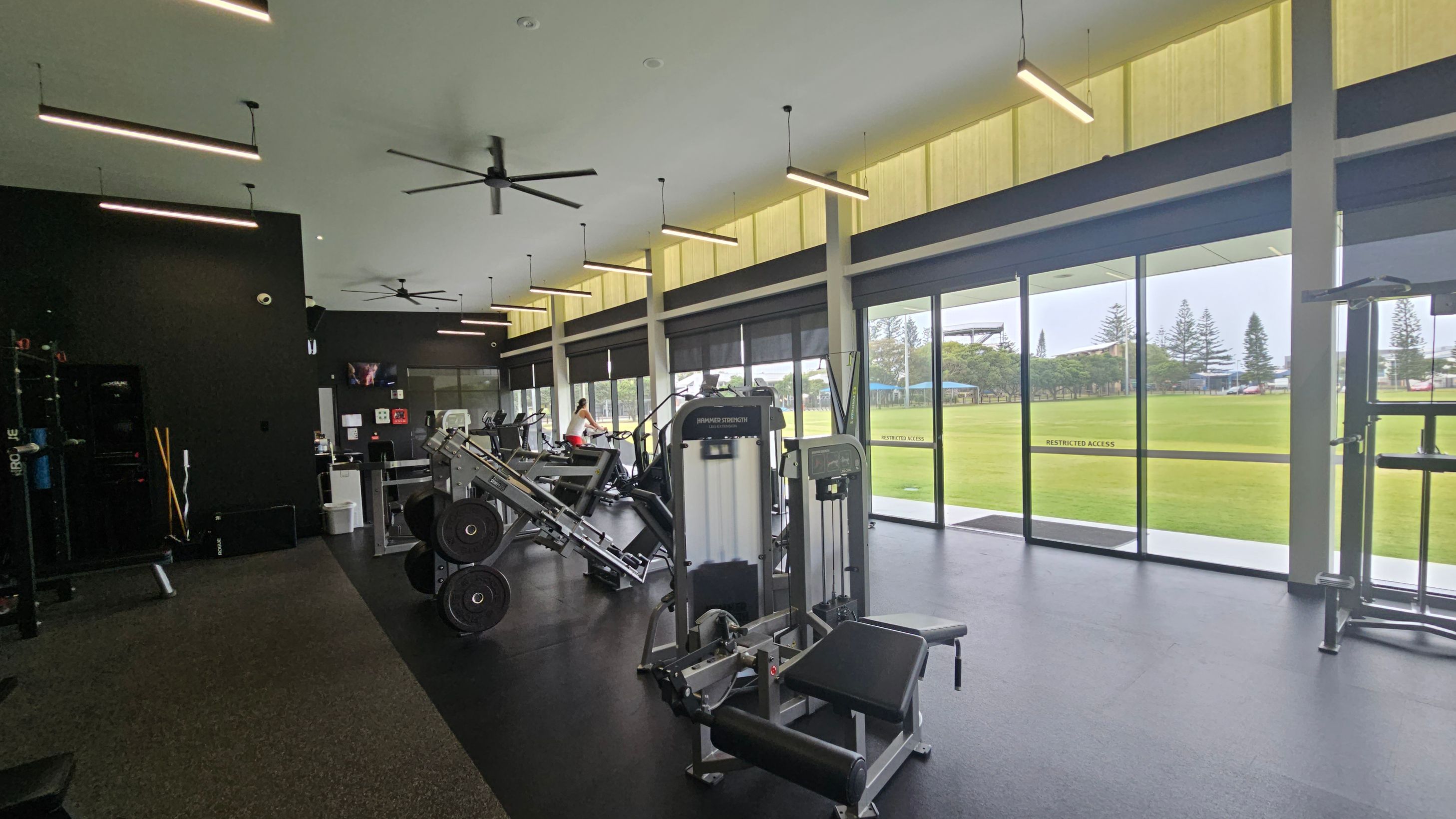 A modern gym interior with exercise equipment on the left, including weight machines and a leg press. The floor is dark with a green section. Large windows on the right provide a view of a grassy field and trees outside. The ceiling is high with linear lights and ceiling fans. A person is using a stationary bike in the background. A modern gym interior with exercise equipment on the left, including weight machines and a leg press. The floor is dark with a green section. Large windows on the right provide a view of a grassy field and trees outside. The ceiling is high with linear lights and ceiling fans. A person is using a stationary bike in the background.