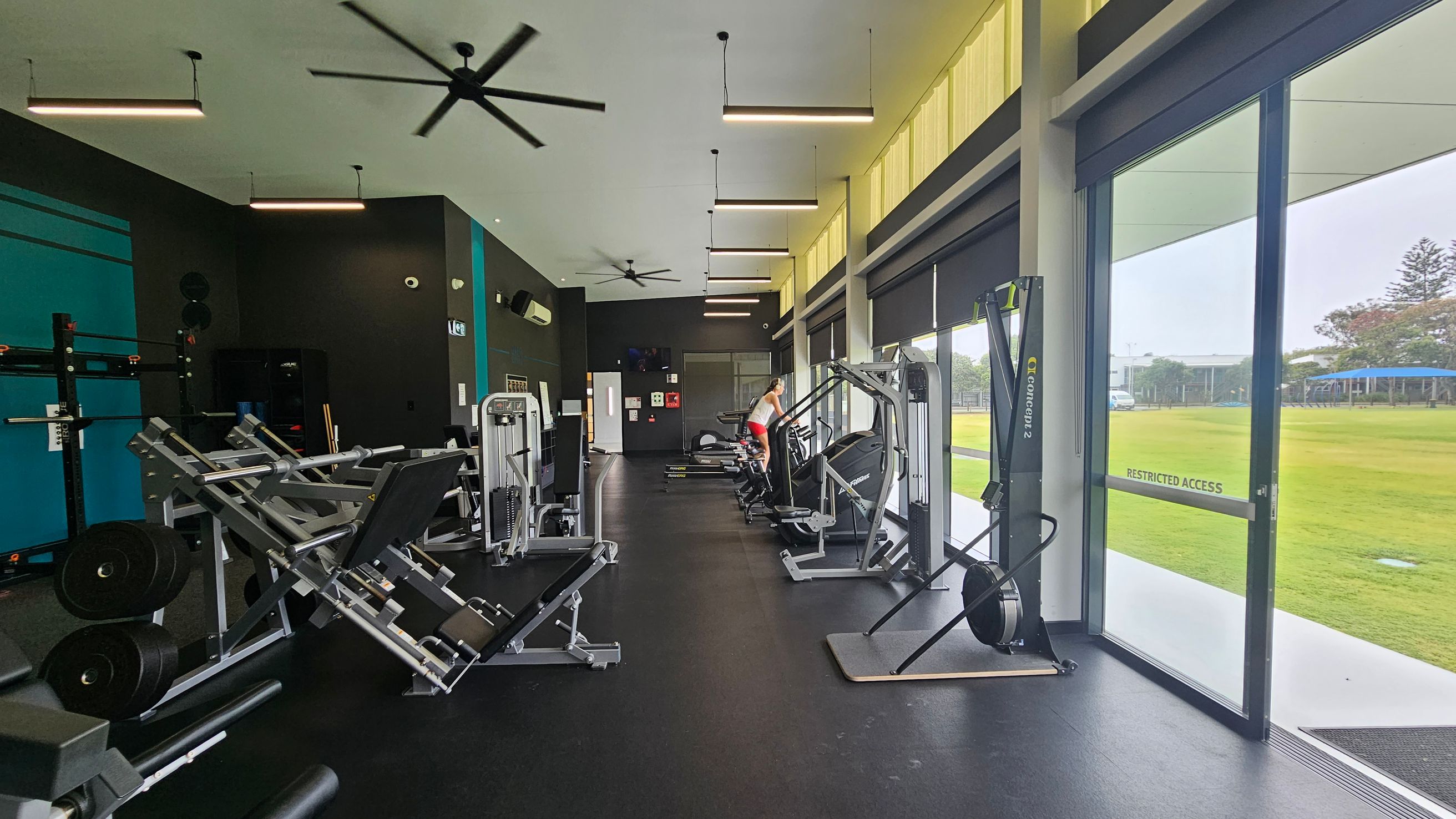 A gym interior with various exercise equipment. On the left, there are weight machines and racks. In the middle, a person is using a treadmill. On the right, large windows and glass doors provide a view of a grassy outdoor area. The floor is dark, and the ceiling has several hanging lights and ceiling fans. A gym interior with various exercise equipment. On the left, there are weight machines and racks. In the middle, a person is using a treadmill. On the right, large windows and glass doors provide a view of a grassy outdoor area. The floor is dark, and the ceiling has several hanging lights and ceiling fans.