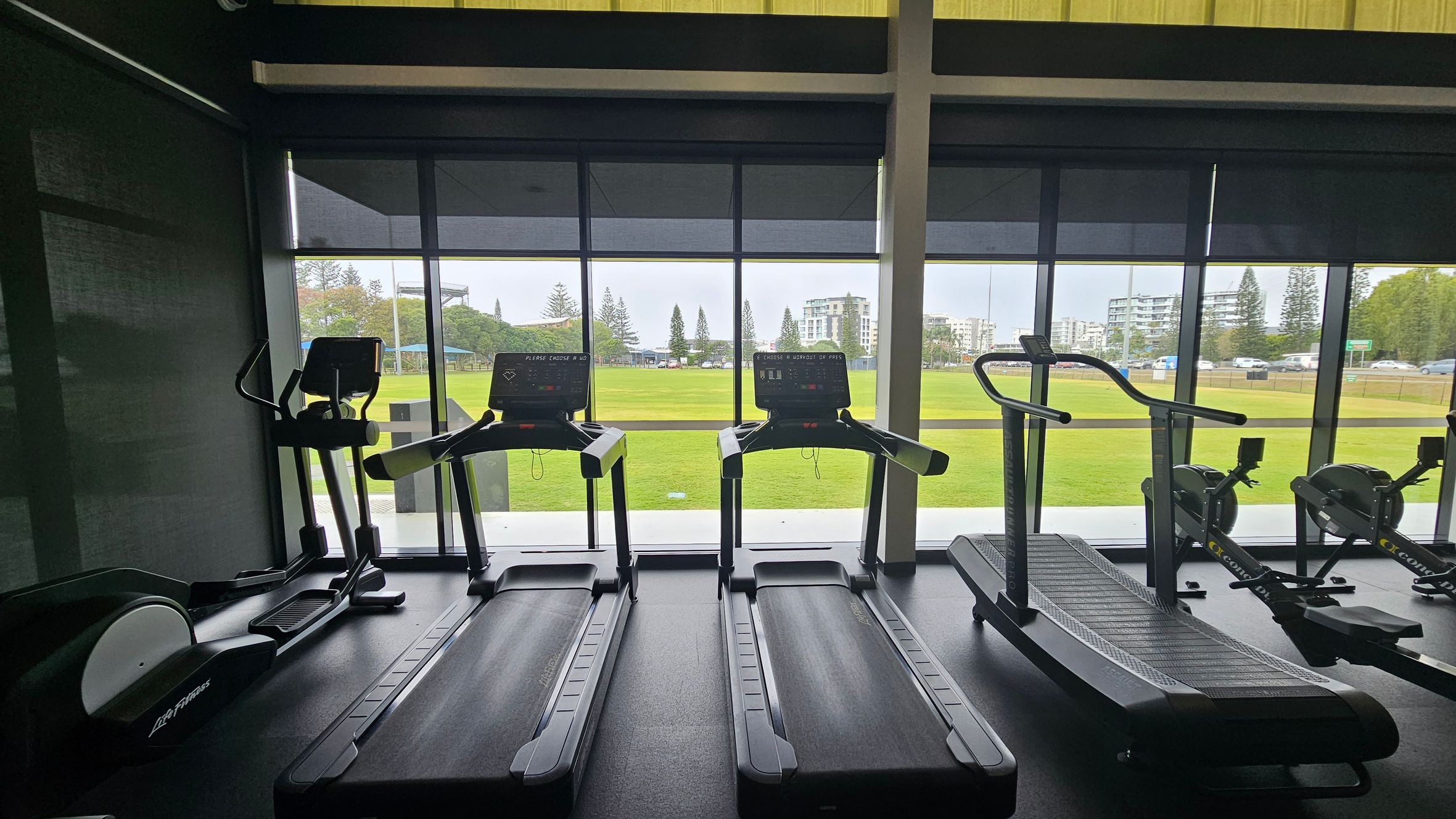 A gym interior with several treadmills and exercise machines lined up in front of large windows. The windows reveal a view of a green field and buildings in the background. The floor is dark and the ceiling is not visible. The equipment includes two standard treadmills, an elliptical machine on the left, and a curved treadmill on the right. The outdoor scene includes trees and a clear sky. A gym interior with several treadmills and exercise machines lined up in front of large windows. The windows reveal a view of a green field and buildings in the background. The floor is dark and the ceiling is not visible. The equipment includes two standard treadmills, an elliptical machine on the left, and a curved treadmill on the right. The outdoor scene includes trees and a clear sky.