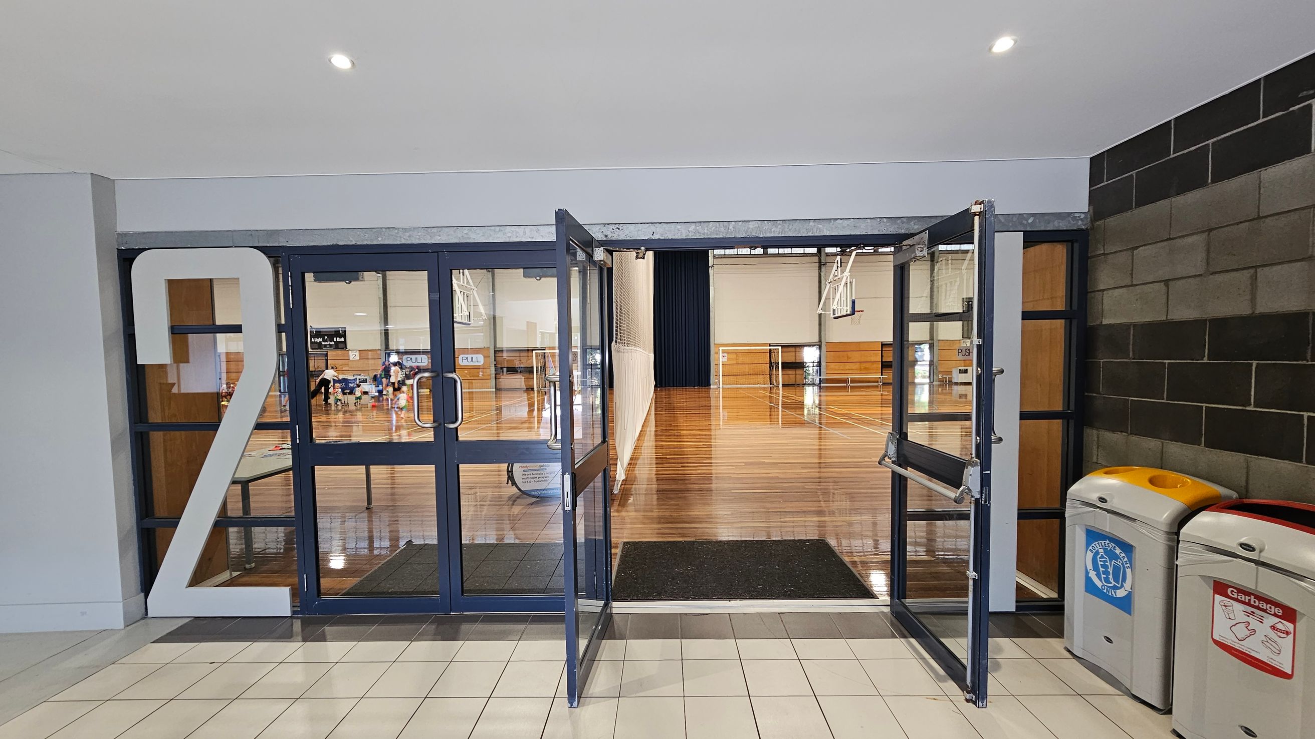 A hallway with a large number "2" on the left side of a glass door entrance. The doors are open, leading into a gymnasium with a polished wooden floor. In the background, there are basketball hoops and a net. On the right side, there are two bins: one for bottles and cans, and another for general garbage. The floor in the foreground is tiled, and the ceiling is plain with recessed lighting. A hallway with a large number "2" on the left side of a glass door entrance. The doors are open, leading into a gymnasium with a polished wooden floor. In the background, there are basketball hoops and a net. On the right side, there are two bins: one for bottles and cans, and another for general garbage. The floor in the foreground is tiled, and the ceiling is plain with recessed lighting.