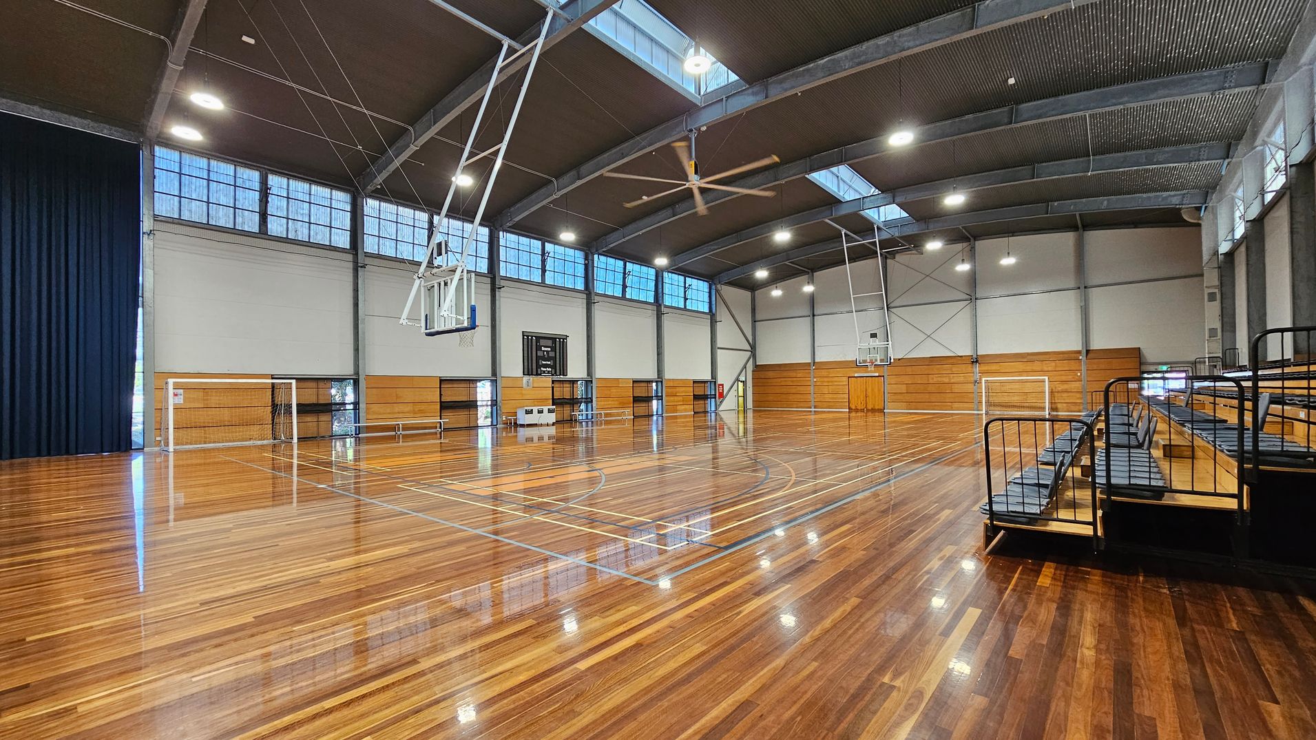 A spacious indoor sports hall with a polished wooden floor. On the left, there is a basketball hoop and a goalpost. The right side features tiered seating with black railings. The ceiling is high with large skylights and industrial lighting. The walls are lined with wooden panels and windows near the top. A scoreboard is visible in the centre background. A spacious indoor sports hall with a polished wooden floor. On the left, there is a basketball hoop and a goalpost. The right side features tiered seating with black railings. The ceiling is high with large skylights and industrial lighting. The walls are lined with wooden panels and windows near the top. A scoreboard is visible in the centre background.