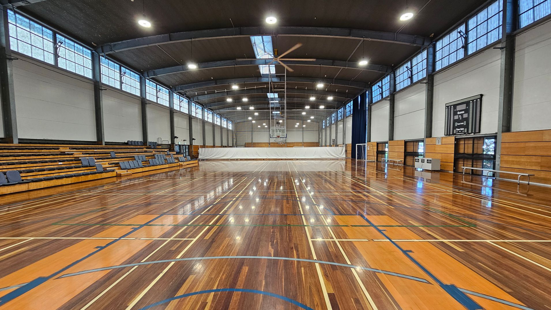 A spacious indoor sports hall with a polished wooden floor marked for various sports. On the left, there are tiered wooden bleachers with blue seats. The right side features a scoreboard and some benches. The ceiling is high with large lights and a central fan. Windows line the upper walls, allowing natural light to enter. A white curtain divides the space at the far end, with a basketball hoop visible above it. A spacious indoor sports hall with a polished wooden floor marked for various sports. On the left, there are tiered wooden bleachers with blue seats. The right side features a scoreboard and some benches. The ceiling is high with large lights and a central fan. Windows line the upper walls, allowing natural light to enter. A white curtain divides the space at the far end, with a basketball hoop visible above it.