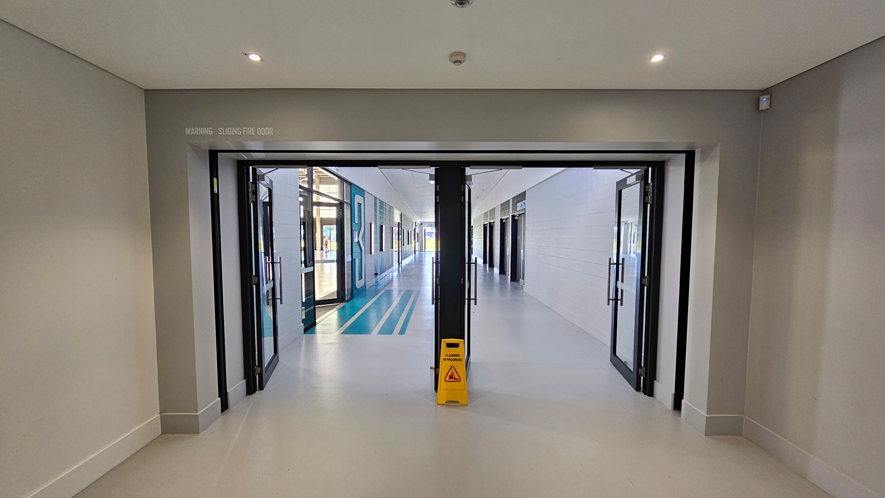 A corridor with a sliding fire door in the centre, partially open, leading to a hallway. The floor is light grey, and a yellow "Caution: Cleaning in Progress" sign is placed in front of the door. The walls are light-coloured, and the ceiling has recessed lighting. The hallway beyond has blue and white striped flooring, with glass doors and windows on the left and white panelled walls on the right. A corridor with a sliding fire door in the centre, partially open, leading to a hallway. The floor is light grey, and a yellow "Caution: Cleaning in Progress" sign is placed in front of the door. The walls are light-coloured, and the ceiling has recessed lighting. The hallway beyond has blue and white striped flooring, with glass doors and windows on the left and white panelled walls on the right.