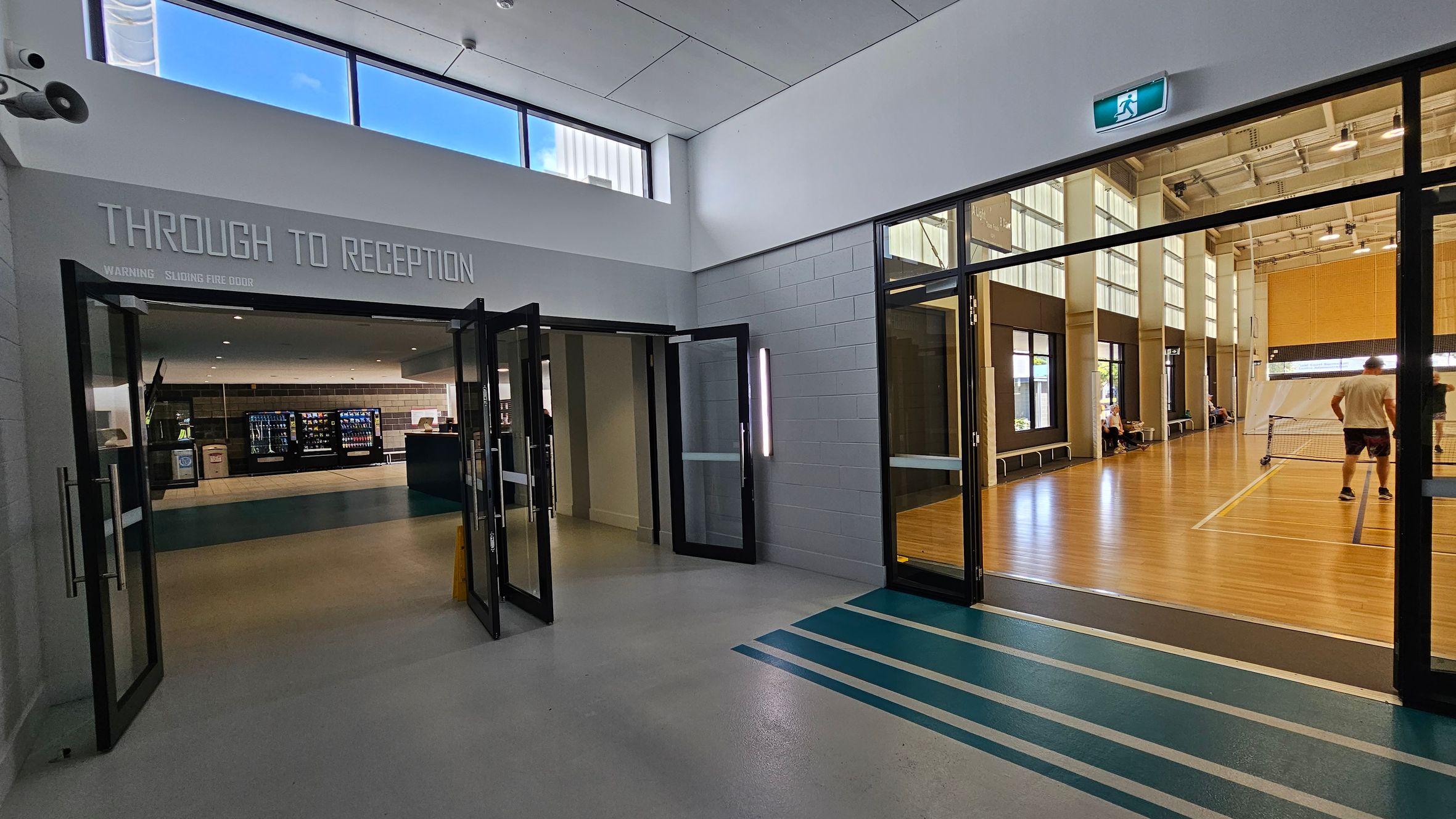 A hallway with a polished floor leads to two areas. On the left, glass doors open to a reception area with vending machines and a counter. On the right, another set of glass doors reveals a sports hall with wooden flooring, where people are playing a game. Above, a high ceiling with windows allows natural light in. An exit sign is visible above the doors to the sports hall. A hallway with a polished floor leads to two areas. On the left, glass doors open to a reception area with vending machines and a counter. On the right, another set of glass doors reveals a sports hall with wooden flooring, where people are playing a game. Above, a high ceiling with windows allows natural light in. An exit sign is visible above the doors to the sports hall.