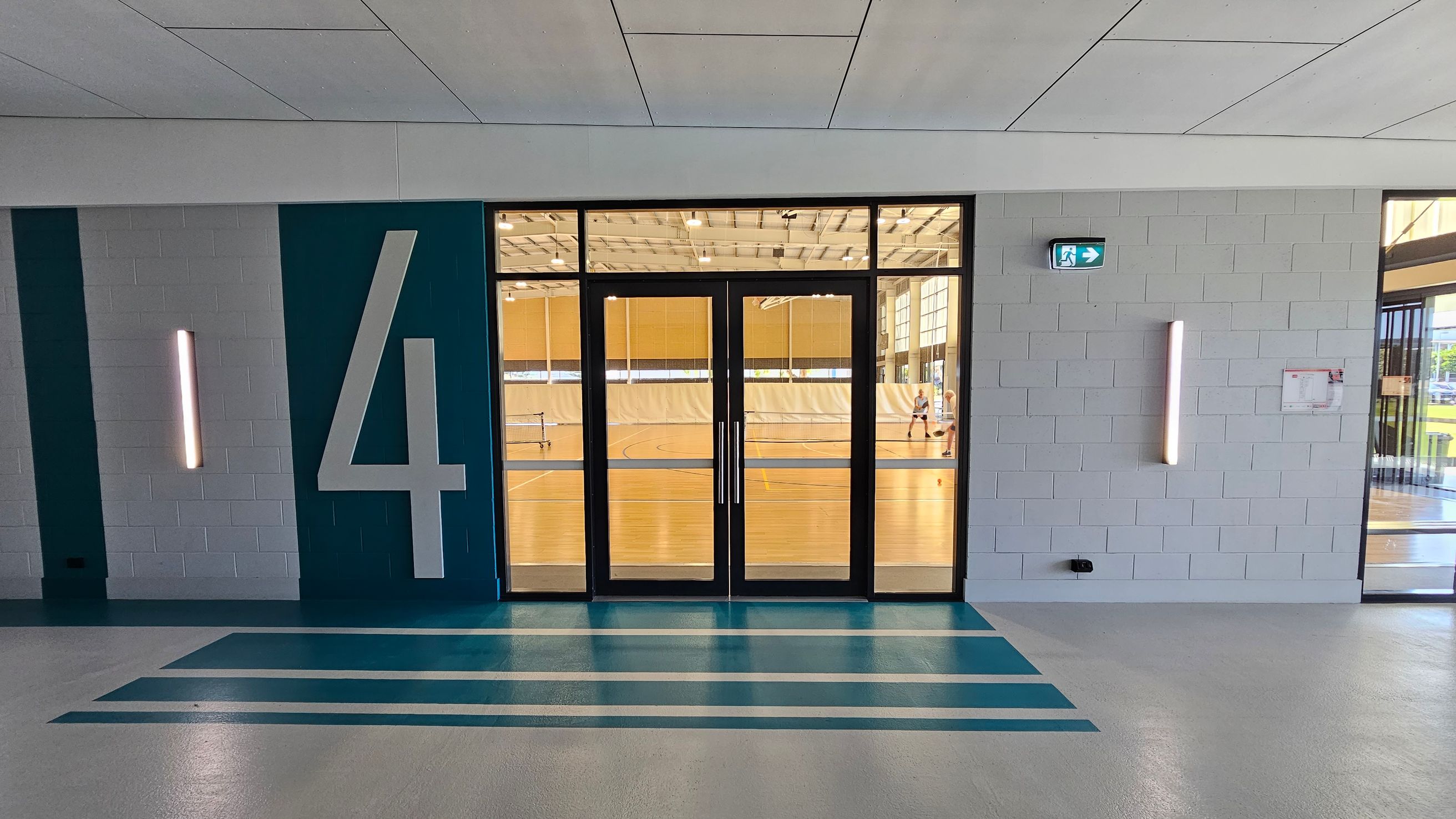 A hallway with a large number "4" on the left wall, painted in white on a teal background. The floor has teal stripes leading to a set of glass double doors. Through the doors, a gymnasium with a wooden floor is visible, where people are playing sports. The walls are light grey with vertical light fixtures on either side of the doors. An emergency exit sign is above the right light fixture. The ceiling is white with rectangular panels. A hallway with a large number "4" on the left wall, painted in white on a teal background. The floor has teal stripes leading to a set of glass double doors. Through the doors, a gymnasium with a wooden floor is visible, where people are playing sports. The walls are light grey with vertical light fixtures on either side of the doors. An emergency exit sign is above the right light fixture. The ceiling is white with rectangular panels.