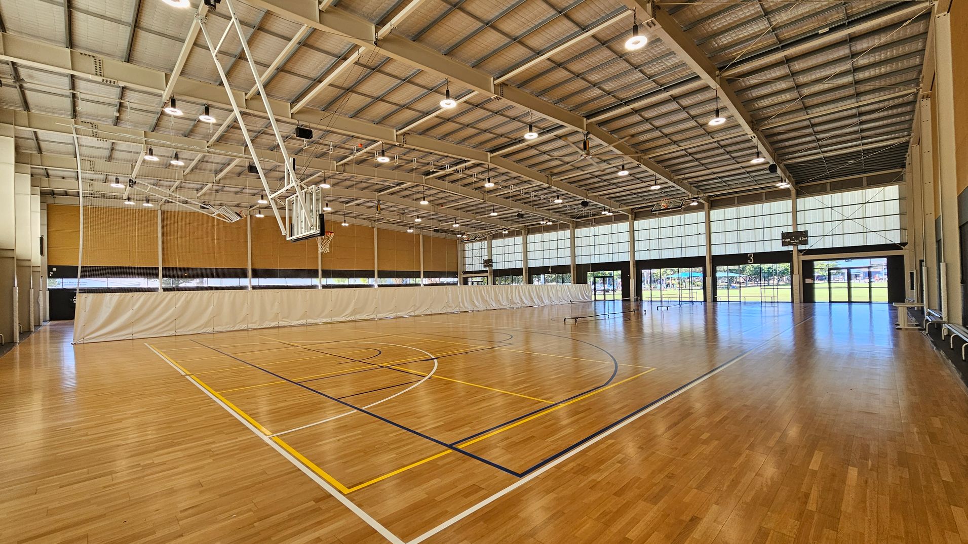 A spacious indoor sports hall with a polished wooden floor marked for basketball. On the left, a basketball hoop is suspended from the ceiling. The ceiling is high with exposed beams and numerous lights. A large white curtain divides the space in the middle. On the right, there are large windows and glass doors allowing natural light to enter, with a view of a grassy area outside. The walls are lined with light-coloured panels. A spacious indoor sports hall with a polished wooden floor marked for basketball. On the left, a basketball hoop is suspended from the ceiling. The ceiling is high with exposed beams and numerous lights. A large white curtain divides the space in the middle. On the right, there are large windows and glass doors allowing natural light to enter, with a view of a grassy area outside. The walls are lined with light-coloured panels.