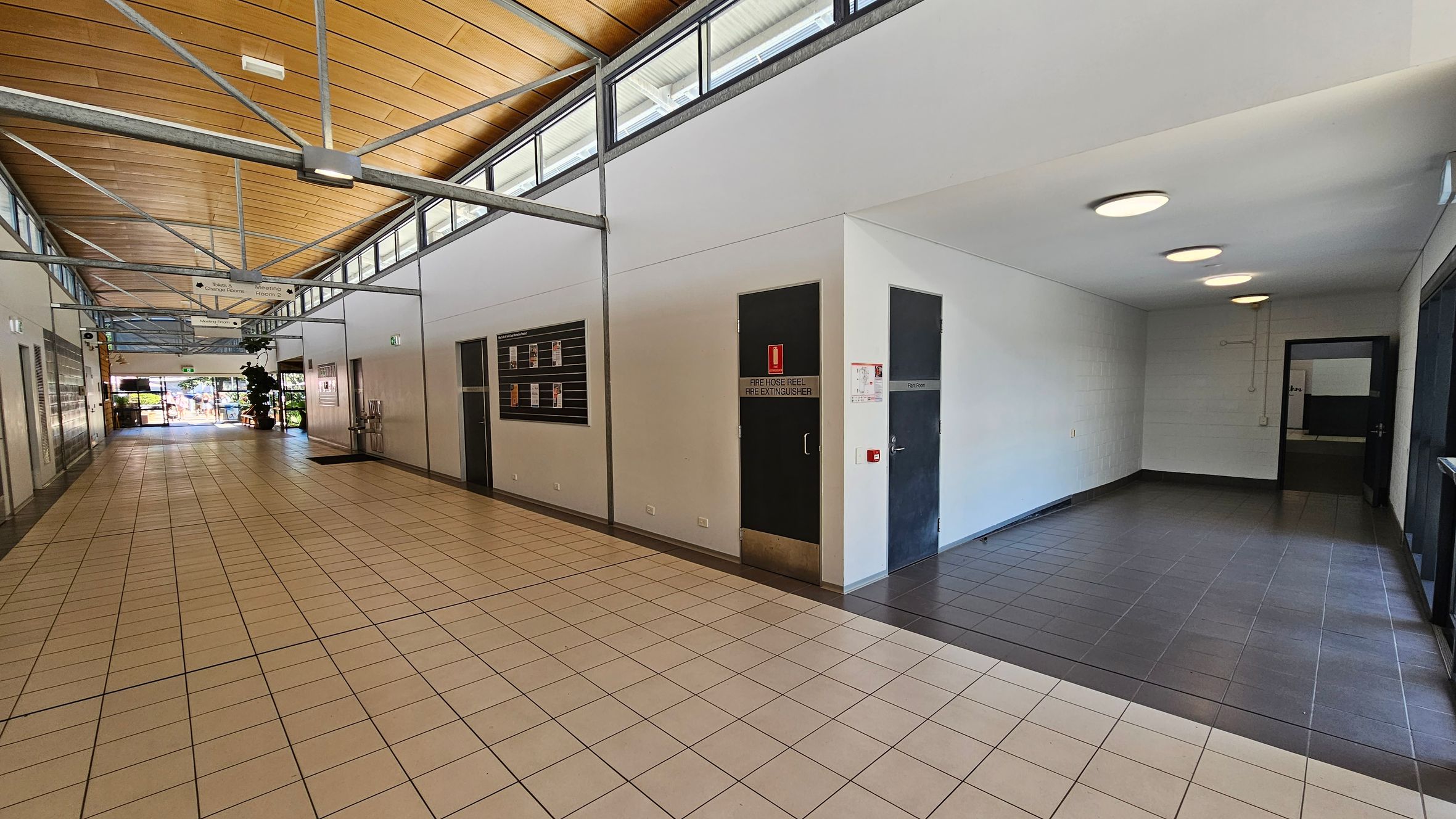 A hallway with a light brown tiled floor and a wooden panelled ceiling. On the left, there are doors and a notice board on the wall. The right side features a dark door labelled "Fire Hose Reel Fire Extinguisher" and another door marked "Plant Room". The ceiling has exposed metal beams and circular lights. At the end of the hallway, there is a glass door leading outside, with some greenery visible through it. A hallway with a light brown tiled floor and a wooden panelled ceiling. On the left, there are doors and a notice board on the wall. The right side features a dark door labelled "Fire Hose Reel Fire Extinguisher" and another door marked "Plant Room". The ceiling has exposed metal beams and circular lights. At the end of the hallway, there is a glass door leading outside, with some greenery visible through it.