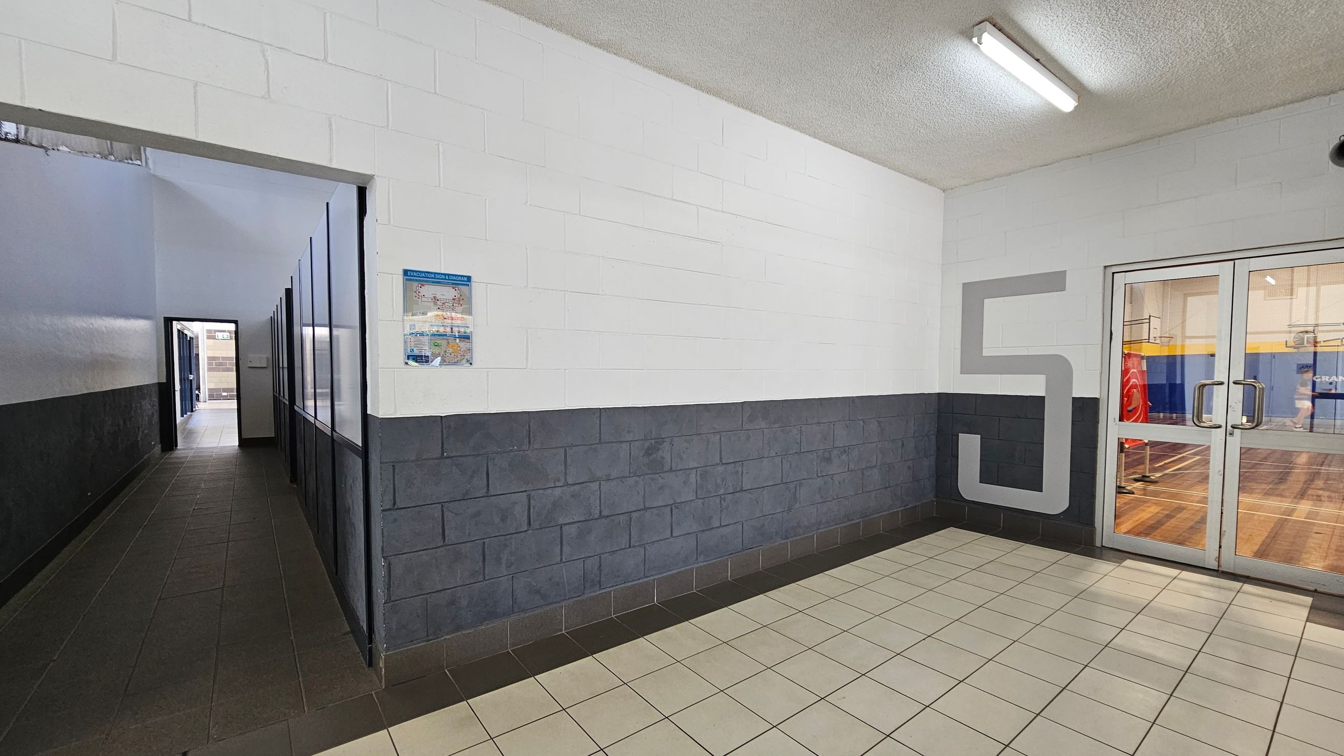 A hallway with a tiled floor and a white ceiling. On the left, a narrow corridor with dark tiles and a series of windows or panels along the wall. On the right, a large number "5" is painted on the wall next to a set of glass double doors leading to a gymnasium with a wooden floor. A poster is mounted on the wall near the corridor entrance. The walls are painted white with a dark grey lower section. Fluorescent lights are mounted on the ceiling. A hallway with a tiled floor and a white ceiling. On the left, a narrow corridor with dark tiles and a series of windows or panels along the wall. On the right, a large number "5" is painted on the wall next to a set of glass double doors leading to a gymnasium with a wooden floor. A poster is mounted on the wall near the corridor entrance. The walls are painted white with a dark grey lower section. Fluorescent lights are mounted on the ceiling.