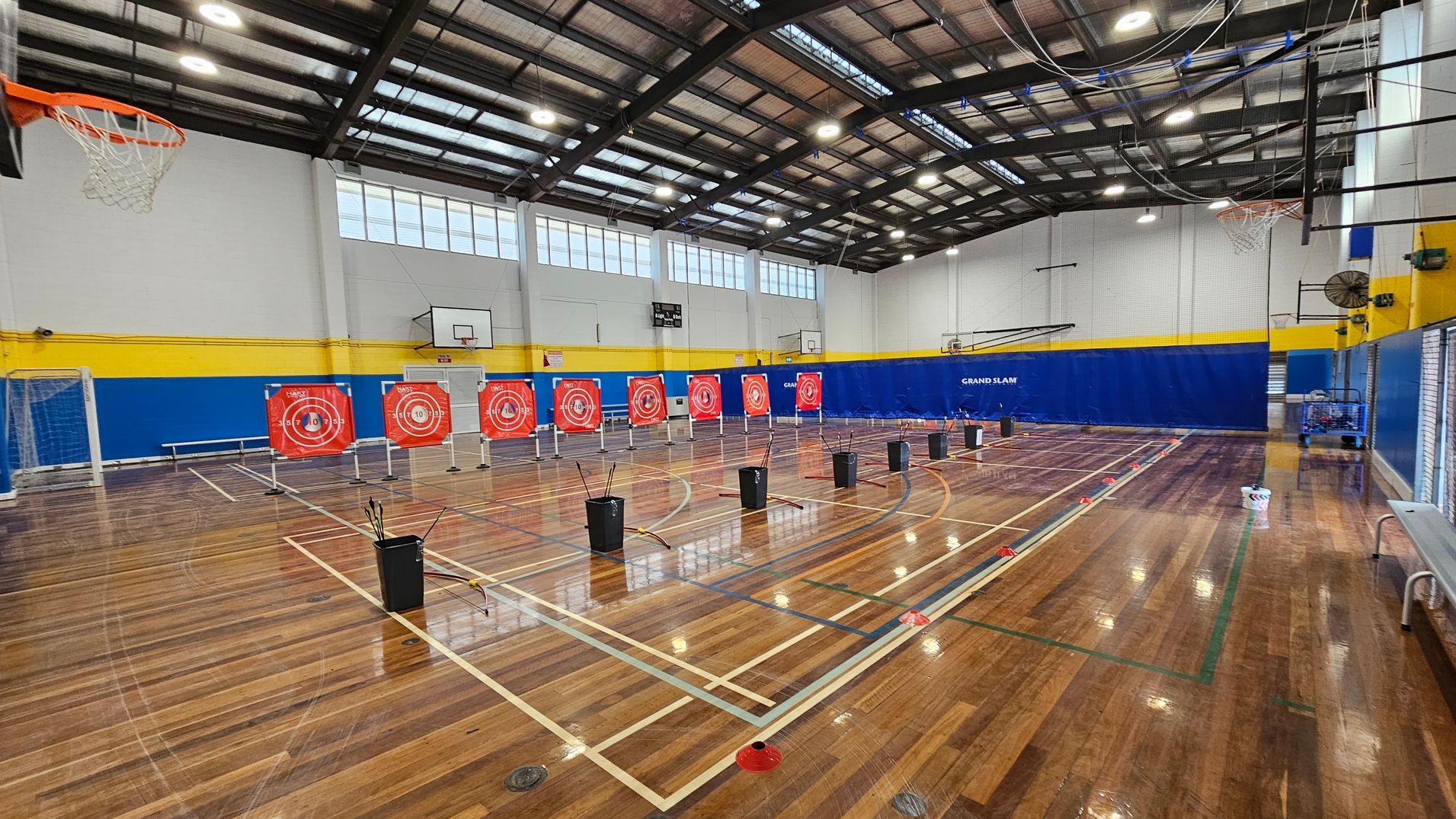 A gymnasium with a polished wooden floor and a high, metal-beamed ceiling. In the centre, there are several archery targets with red and white concentric circles. Black bins with arrows are placed in front of each target. On the left, a basketball hoop and a goalpost are visible. The walls are painted white with a blue and yellow stripe running horizontally. On the right, a bench and a blue wall with "GRAND SLAM" written on it can be seen. A gymnasium with a polished wooden floor and a high, metal-beamed ceiling. In the centre, there are several archery targets with red and white concentric circles. Black bins with arrows are placed in front of each target. On the left, a basketball hoop and a goalpost are visible. The walls are painted white with a blue and yellow stripe running horizontally. On the right, a bench and a blue wall with "GRAND SLAM" written on it can be seen.