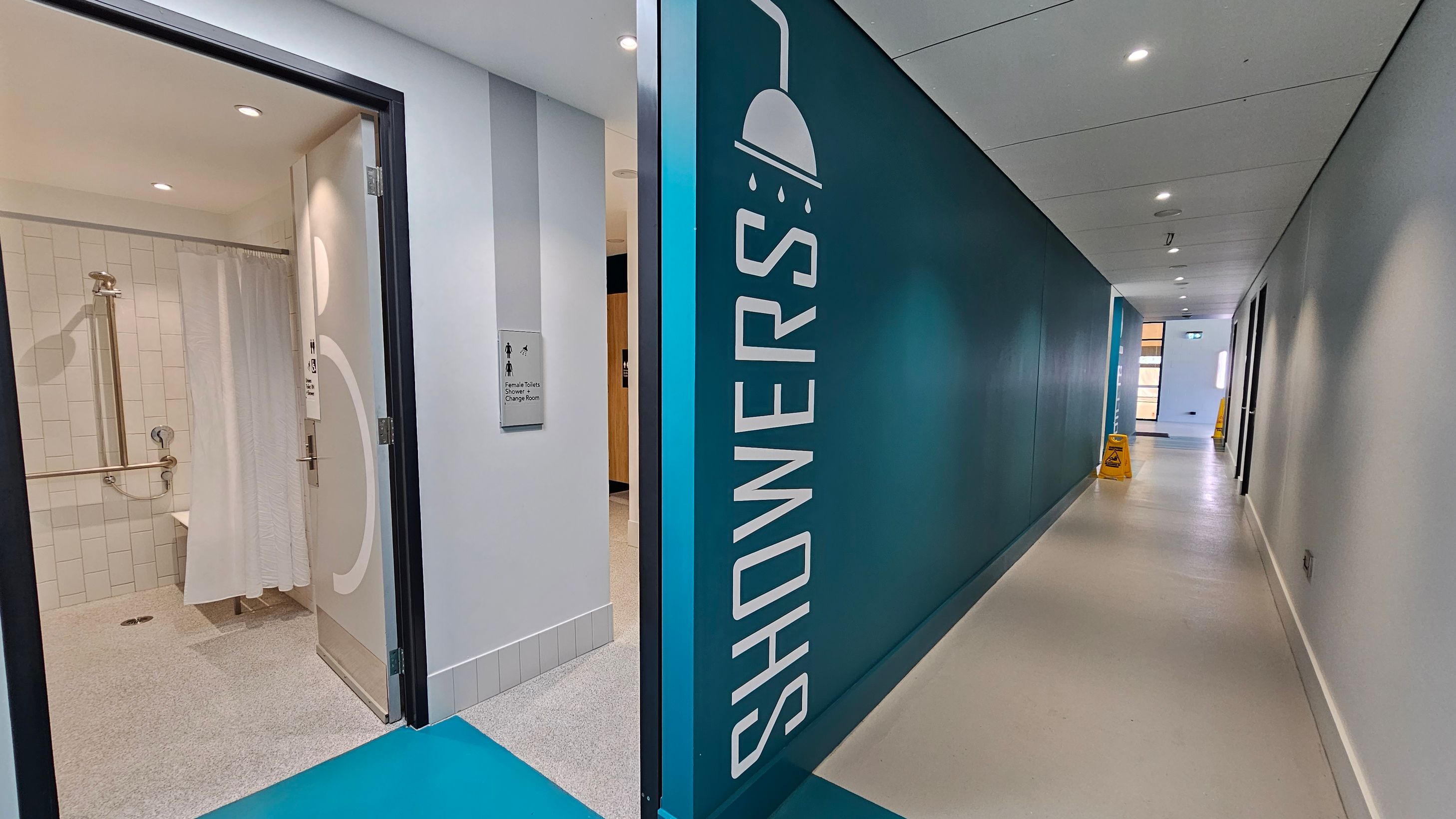 A corridor with a teal wall on the right featuring a large "SHOWERS" sign and a showerhead icon. On the left, an open door leads to a shower room with white tiles and a shower curtain. A sign next to the door reads "Female Toilets Change Room." The floor is light-coloured, and the ceiling has recessed lighting. Further down the corridor, a yellow caution sign is visible near a windowed area. A corridor with a teal wall on the right featuring a large "SHOWERS" sign and a showerhead icon. On the left, an open door leads to a shower room with white tiles and a shower curtain. A sign next to the door reads "Female Toilets Change Room." The floor is light-coloured, and the ceiling has recessed lighting. Further down the corridor, a yellow caution sign is visible near a windowed area.