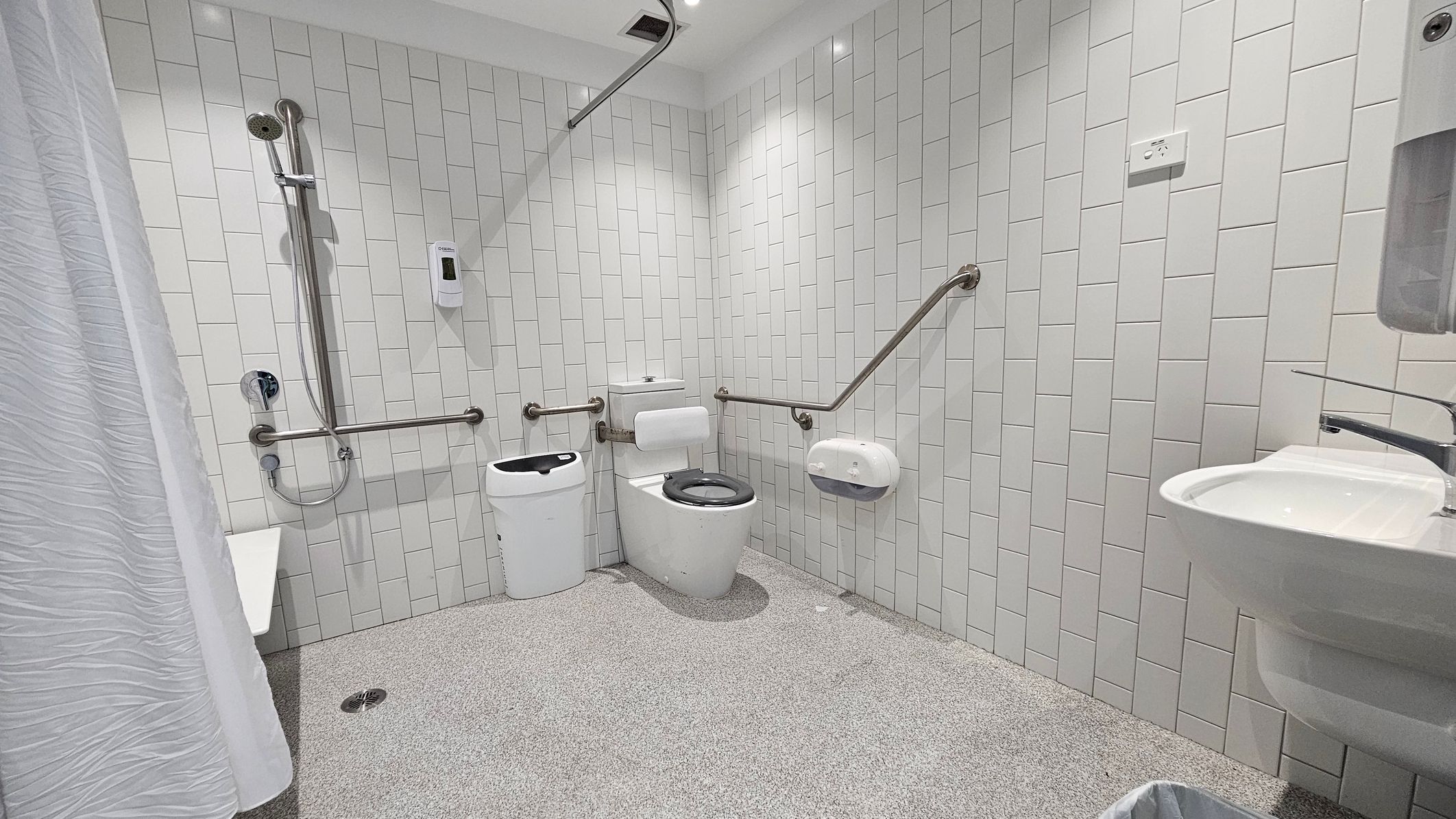 A modern, accessible bathroom with white tiled walls and a speckled grey floor. On the left, there's a shower area with a handheld showerhead and a white curtain. In the centre, a toilet with a black seat is flanked by grab bars. A sanitary bin is next to the toilet. On the right, a wall-mounted sink with a soap dispenser above it. The ceiling is white with recessed lighting. A modern, accessible bathroom with white tiled walls and a speckled grey floor. On the left, there's a shower area with a handheld showerhead and a white curtain. In the centre, a toilet with a black seat is flanked by grab bars. A sanitary bin is next to the toilet. On the right, a wall-mounted sink with a soap dispenser above it. The ceiling is white with recessed lighting.