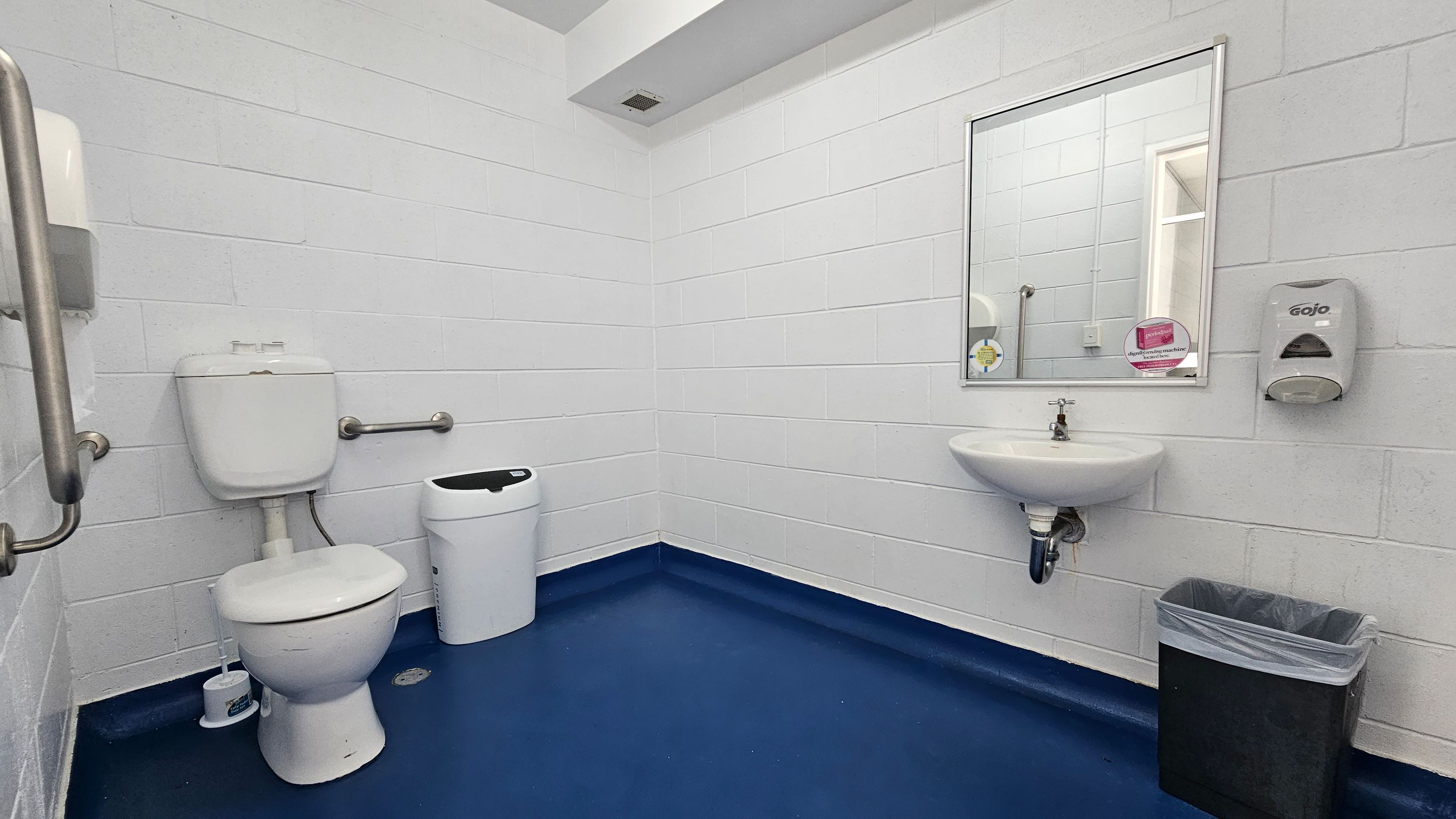 A public bathroom with white brick walls and a blue floor. On the left, there is a toilet with a grab bar and a small bin nearby. In the centre, a wall-mounted sink is below a rectangular mirror. To the right of the sink, a soap dispenser is mounted on the wall. Below the dispenser, there is a black waste bin with a liner. The ceiling is white with a small vent. A public bathroom with white brick walls and a blue floor. On the left, there is a toilet with a grab bar and a small bin nearby. In the centre, a wall-mounted sink is below a rectangular mirror. To the right of the sink, a soap dispenser is mounted on the wall. Below the dispenser, there is a black waste bin with a liner. The ceiling is white with a small vent.