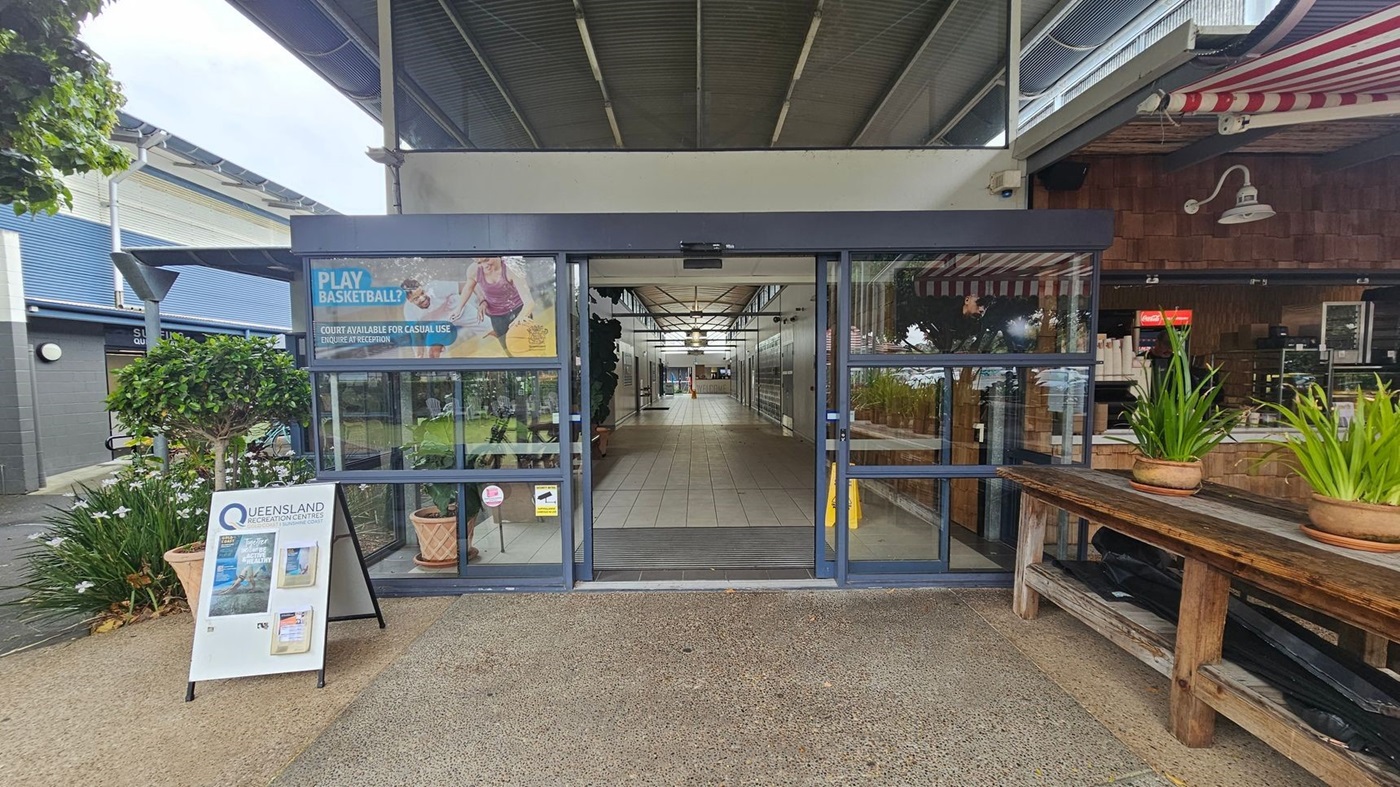 Entrance to a building with glass doors in the centre. A sign on the left reads "Queensland Recreation Centres" with promotional material. A small tree and flowers are nearby. On the right, a wooden bench holds potted plants, and a café with a red and white striped awning is visible. The floor is tiled, and the ceiling has exposed beams and lights. Entrance to a building with glass doors in the centre. A sign on the left reads "Queensland Recreation Centres" with promotional material. A small tree and flowers are nearby. On the right, a wooden bench holds potted plants, and a café with a red and white striped awning is visible. The floor is tiled, and the ceiling has exposed beams and lights.
