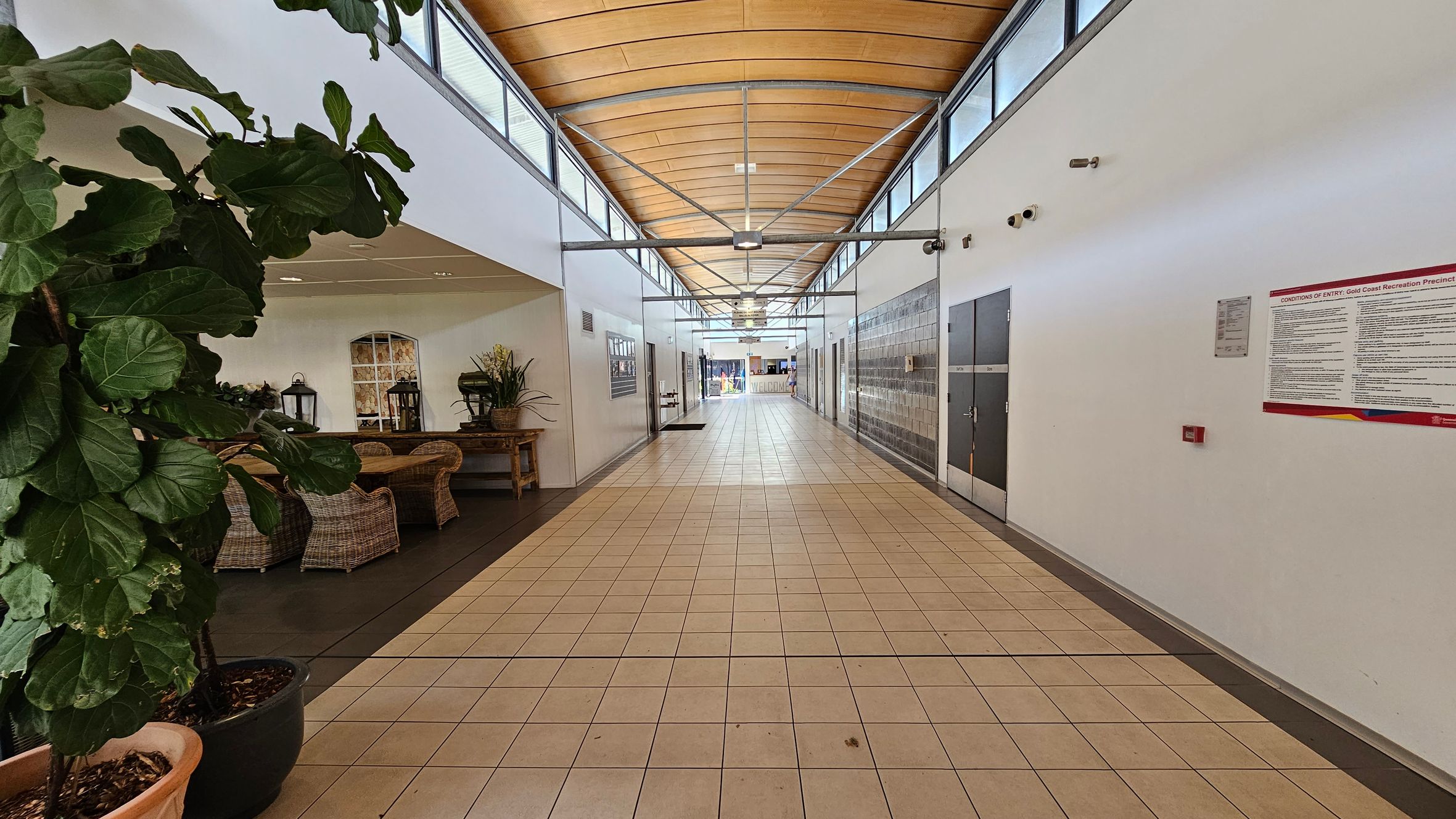 A corridor with a tiled floor and a wooden panelled ceiling. On the left, there are large potted plants and wicker furniture, including chairs and a table with decorative items. The right side features a white wall with a notice board and security cameras. The corridor extends into the distance, with doors and windows along the sides, leading to a bright area at the end. A corridor with a tiled floor and a wooden panelled ceiling. On the left, there are large potted plants and wicker furniture, including chairs and a table with decorative items. The right side features a white wall with a notice board and security cameras. The corridor extends into the distance, with doors and windows along the sides, leading to a bright area at the end.