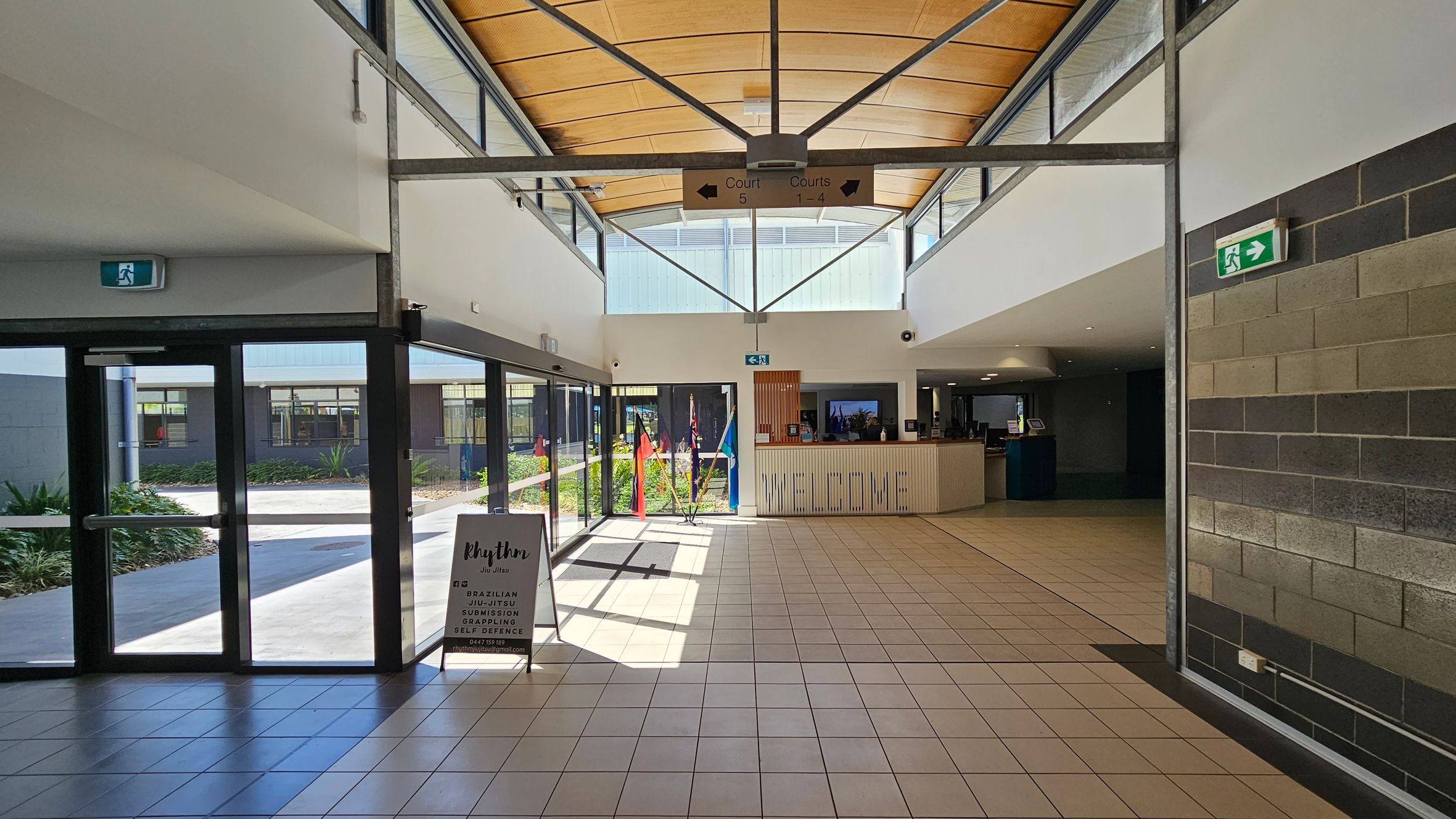 A spacious indoor area with a tiled floor and a high, wooden ceiling. On the left, there are glass doors leading outside, with a sign advertising "Rhythm Jiu Jitsu" nearby. In the centre, a reception desk with a "WELCOME" sign is visible, and flags are displayed behind it. On the right, a wall with grey and brown bricks. Above, a sign directs to courts 1-5. Emergency exit signs are positioned on both sides. Natural light streams in through large windows. A spacious indoor area with a tiled floor and a high, wooden ceiling. On the left, there are glass doors leading outside, with a sign advertising "Rhythm Jiu Jitsu" nearby. In the centre, a reception desk with a "WELCOME" sign is visible, and flags are displayed behind it. On the right, a wall with grey and brown bricks. Above, a sign directs to courts 1-5. Emergency exit signs are positioned on both sides. Natural light streams in through large windows.