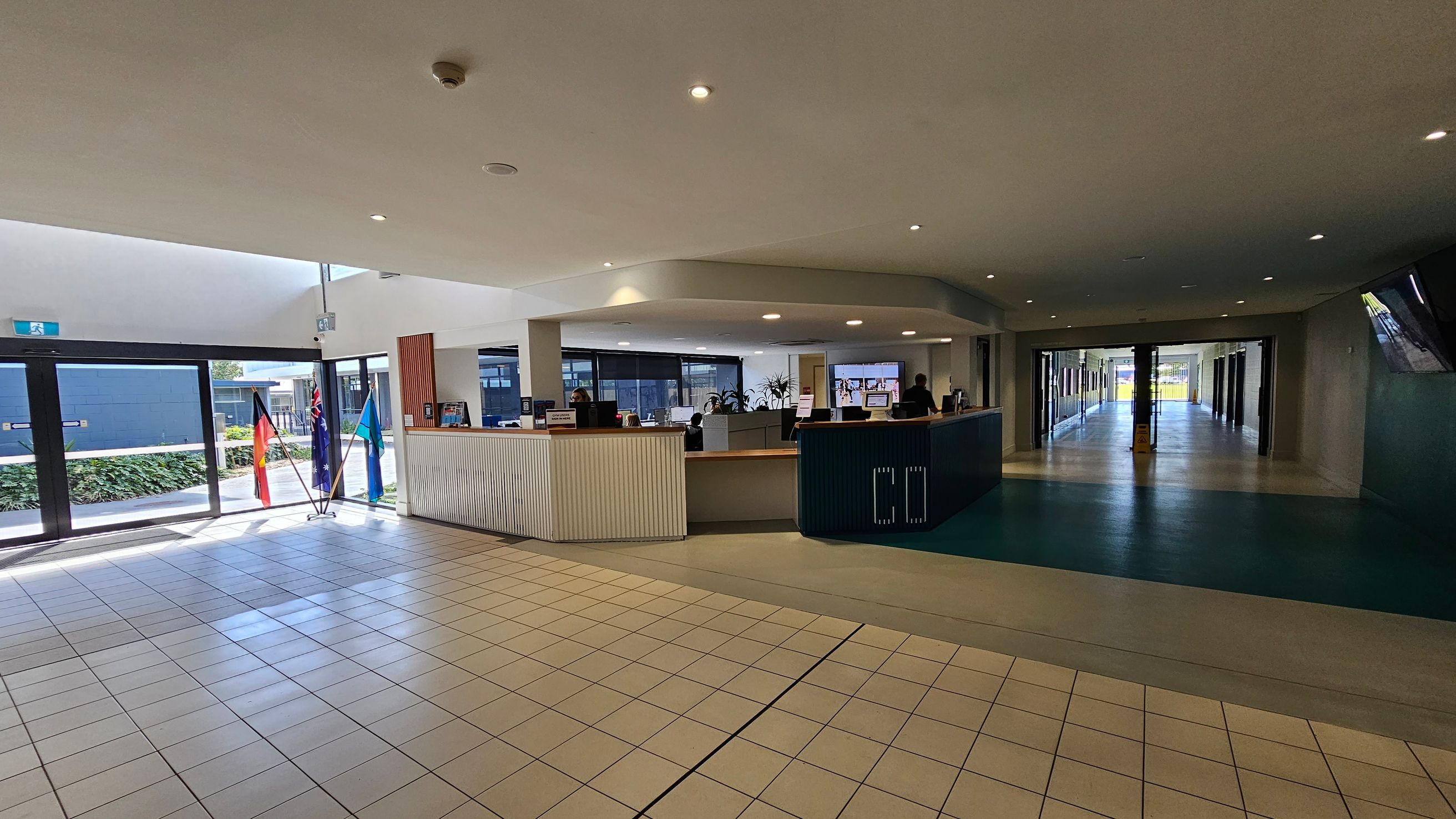 A spacious indoor area with a tiled floor and a high ceiling with recessed lighting. On the left, there are large glass doors with three flags displayed nearby. In the centre, a curved reception desk is staffed by people, with computer monitors and potted plants visible. To the right, a corridor extends into the distance, with a polished floor and glass doors at the end. The area is well-lit with natural light coming through the glass doors. A spacious indoor area with a tiled floor and a high ceiling with recessed lighting. On the left, there are large glass doors with three flags displayed nearby. In the centre, a curved reception desk is staffed by people, with computer monitors and potted plants visible. To the right, a corridor extends into the distance, with a polished floor and glass doors at the end. The area is well-lit with natural light coming through the glass doors.