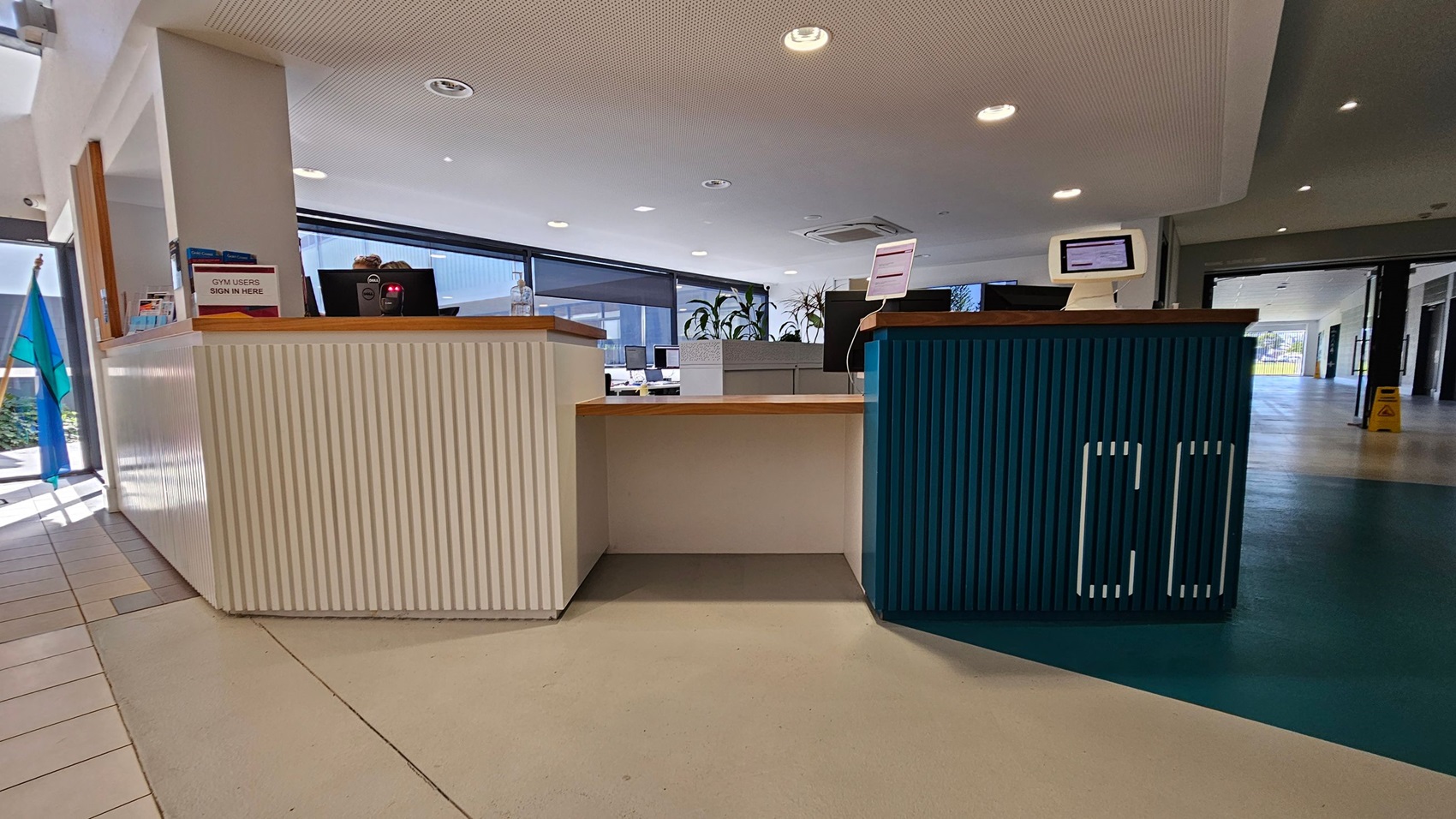 A reception area with a counter in the centre. The left side of the counter is white with a sign that reads "GYM USERS SIGN IN HERE" and some brochures. The right side is blue with a computer monitor. Behind the counter are more monitors and potted plants. The floor is a mix of light and dark tiles, and the ceiling is white with recessed lighting. Large windows in the background let in natural light. A reception area with a counter in the centre. The left side of the counter is white with a sign that reads "GYM USERS SIGN IN HERE" and some brochures. The right side is blue with a computer monitor. Behind the counter are more monitors and potted plants. The floor is a mix of light and dark tiles, and the ceiling is white with recessed lighting. Large windows in the background let in natural light.